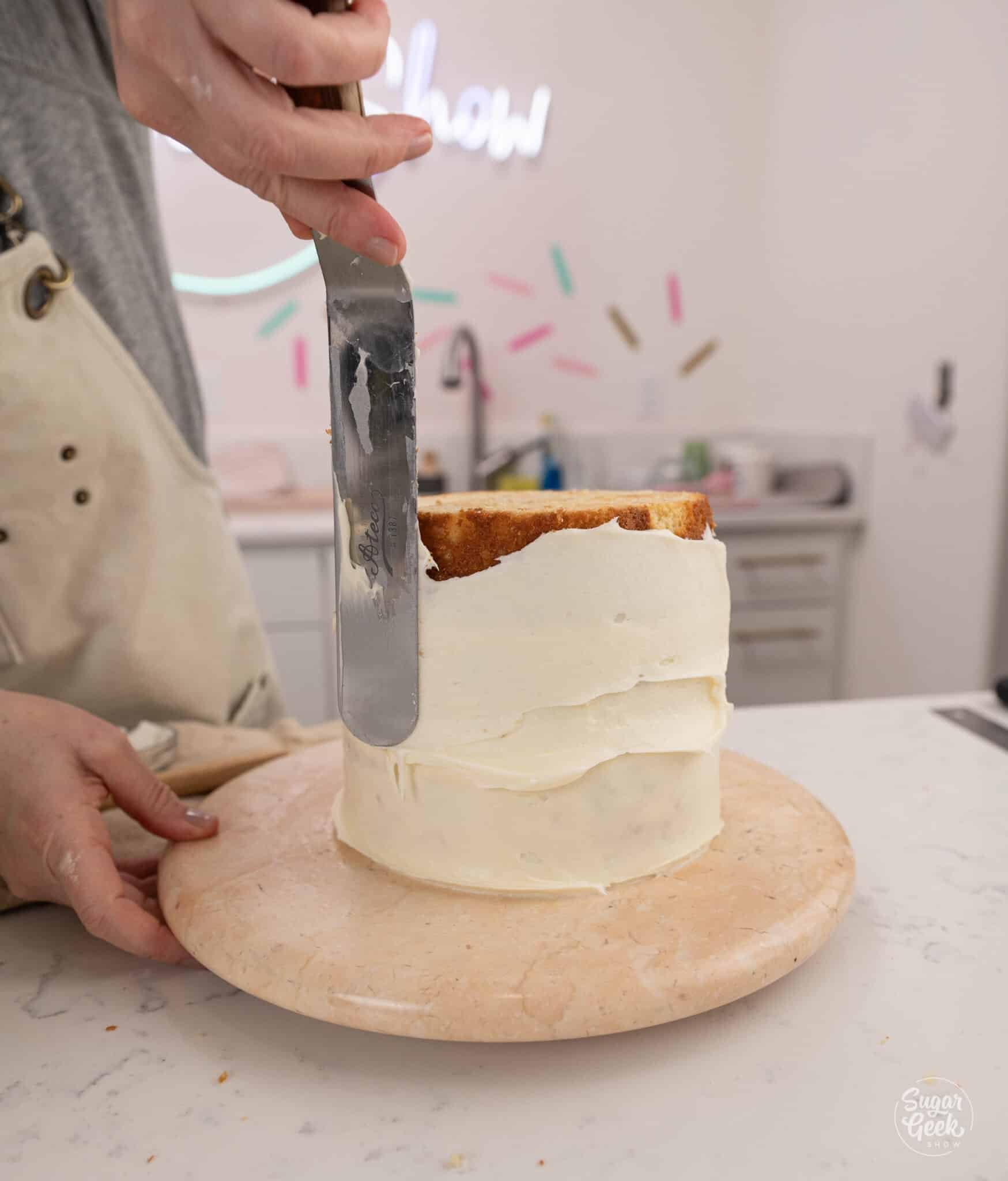hand spreading a thin layer of cream cheese frosting on a cake with an offset spatula