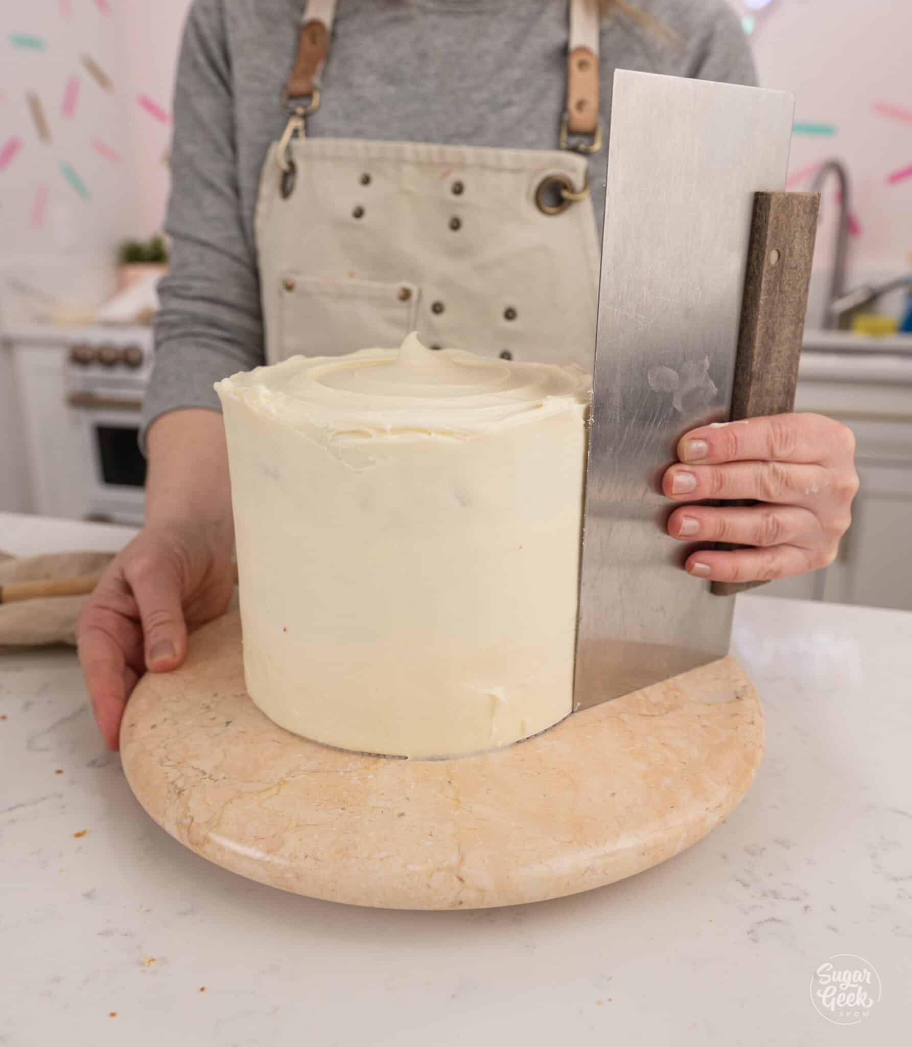 hand smoothing the frosting on the side of a cake with a bench scraper