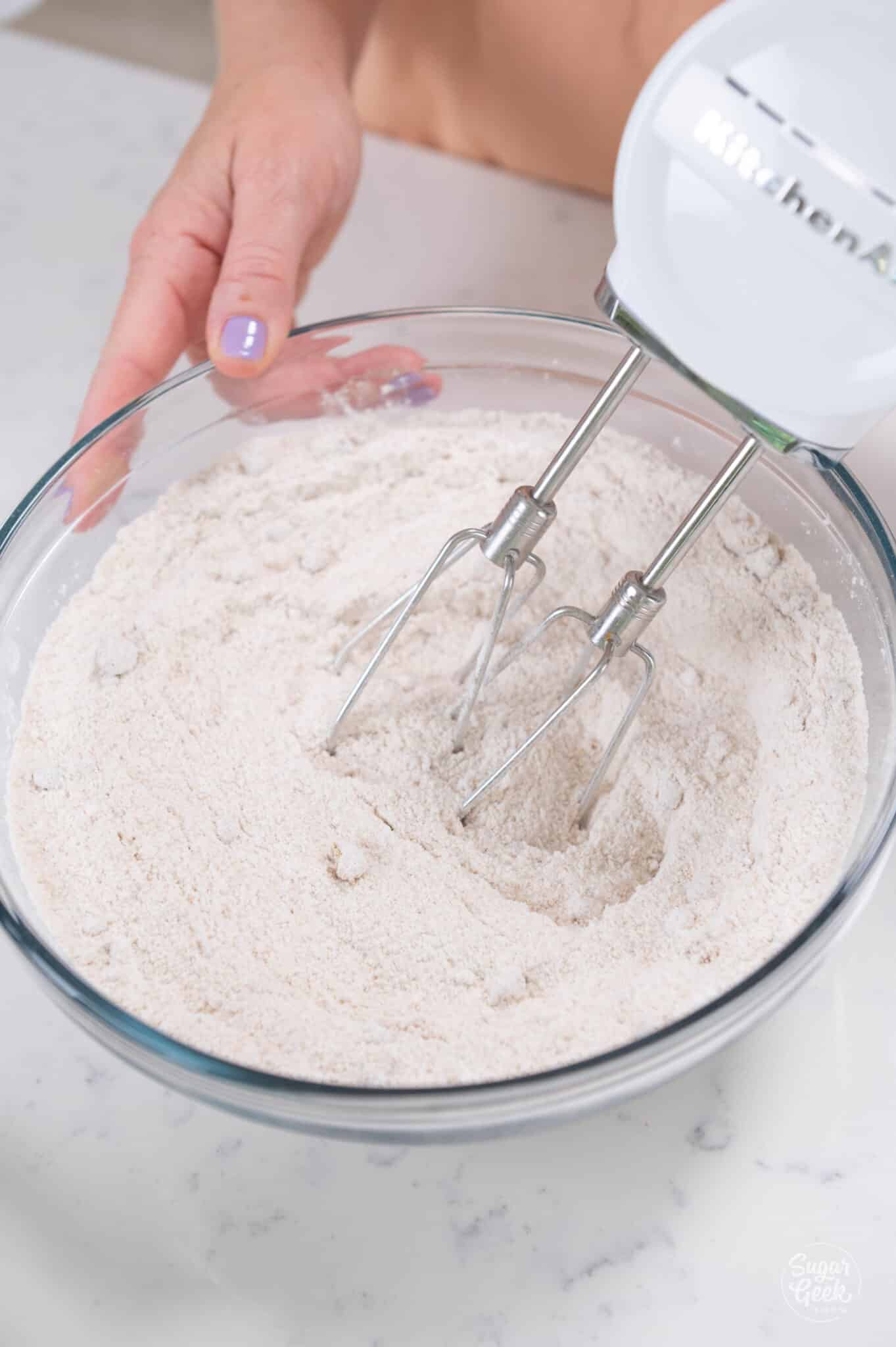 hands using hand mixer to whisk dry ingredients in bowl.