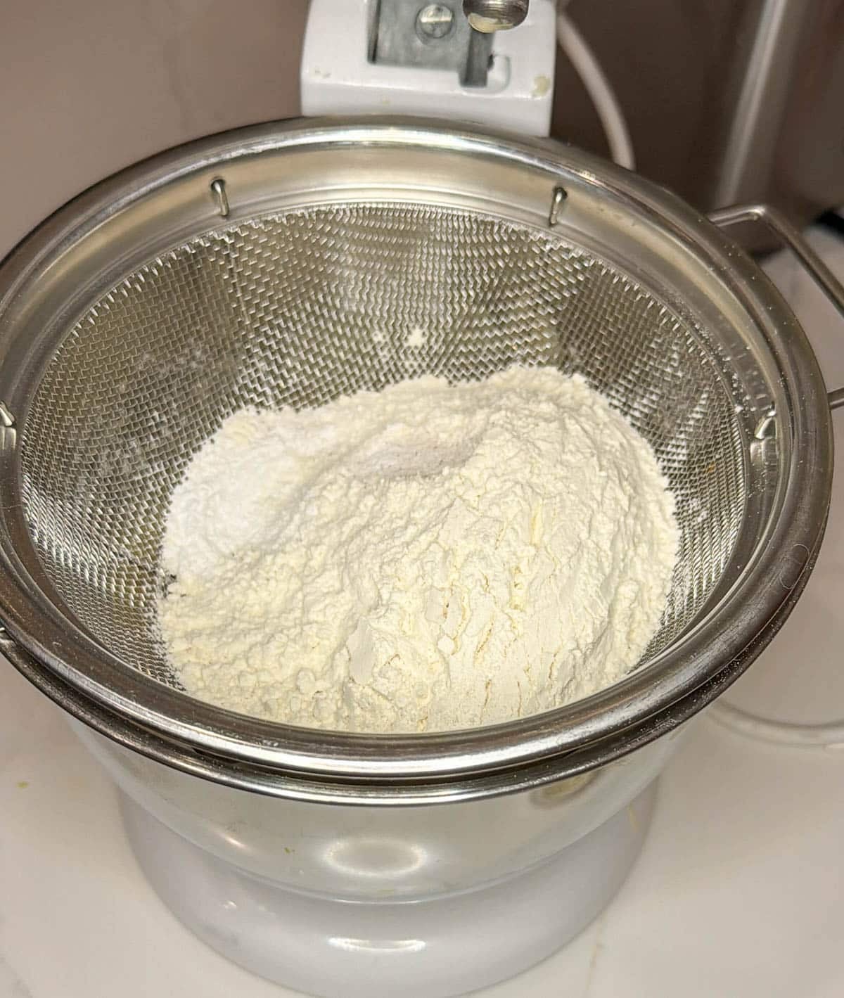 Sifter with flour mixture inside on top of the bowl of a stand mixer.