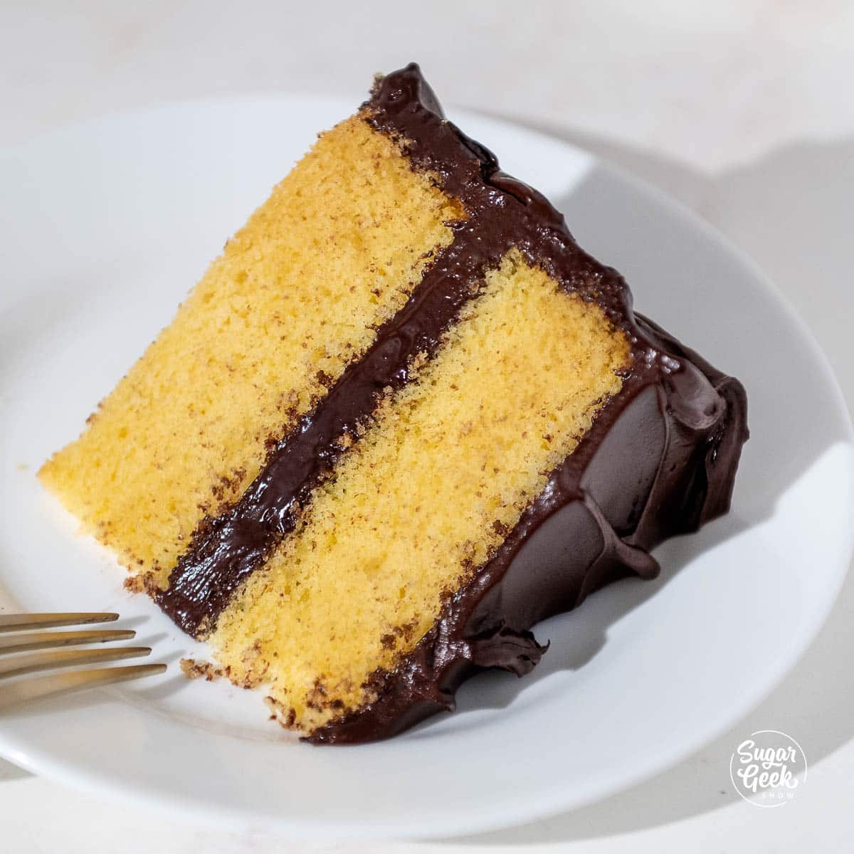 close up of a yellow cake slice with ganache frosting on a white plate