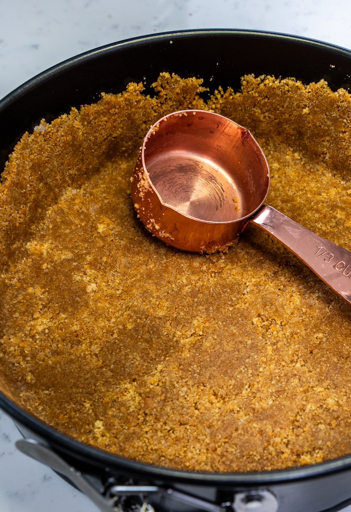 close up of graham cracker mixture in a cheesecake pan with a measuring cup