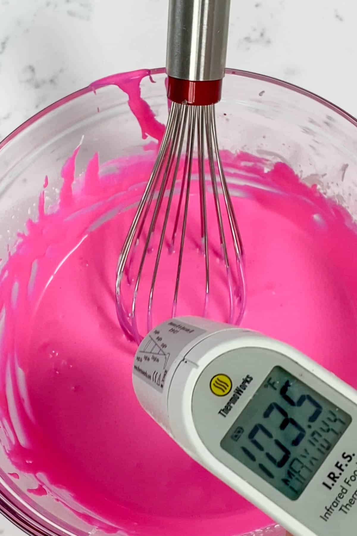 Pink ganache with a whisk in a clear bowl with a laser thermometer above.