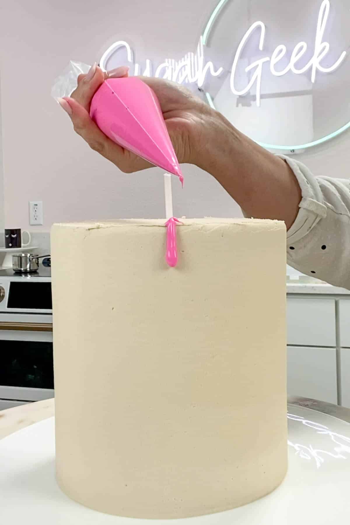 Piping bag with pink ganache piping a drip onto a white cake.