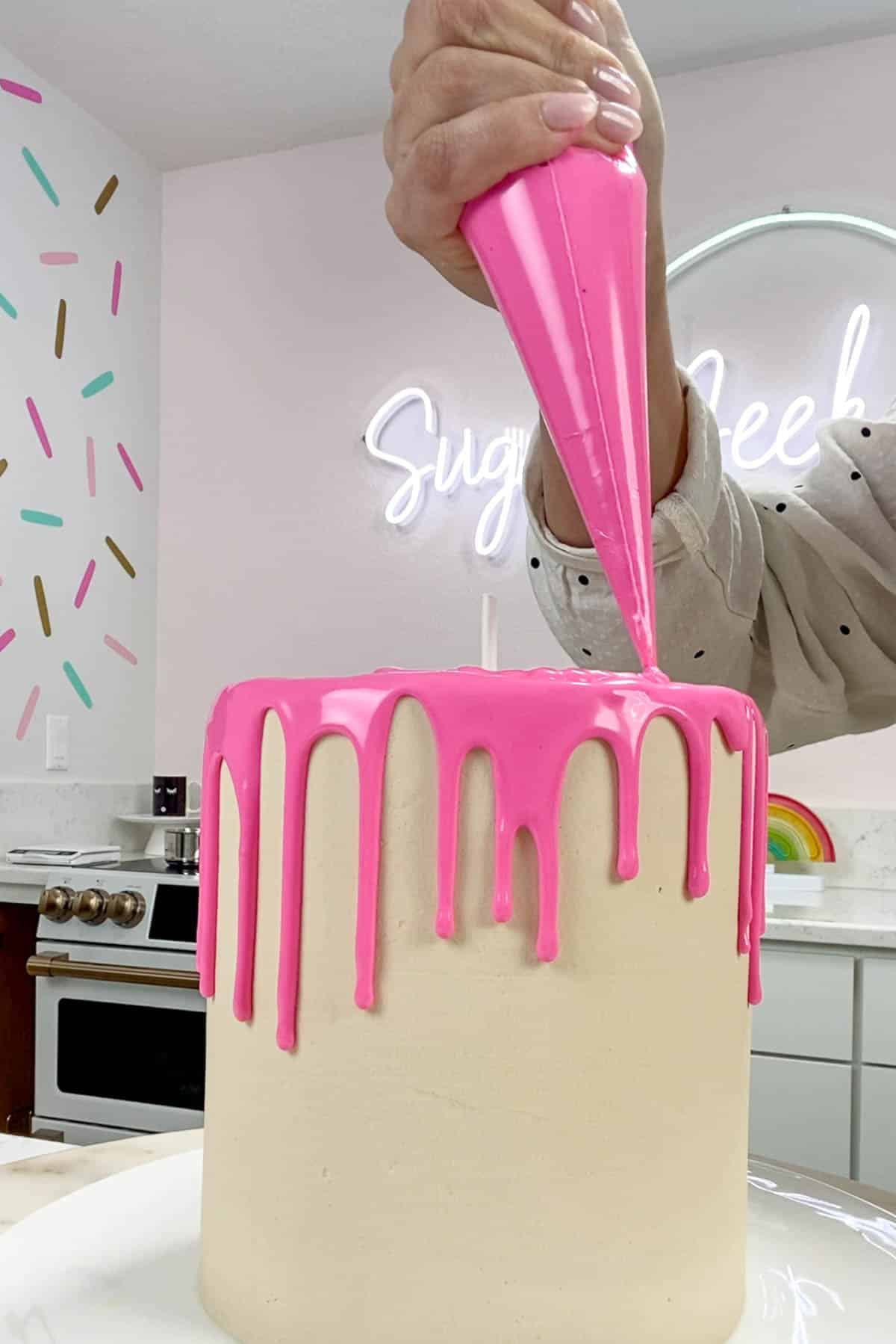 Piping bag piping pink ganache on to the top of a white cake.