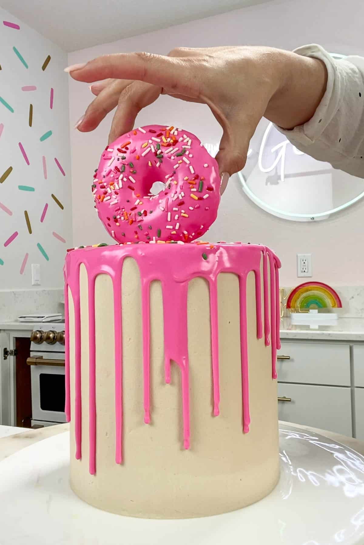 Hand placing a pink donut on top of a white cake with pink ganache drips.