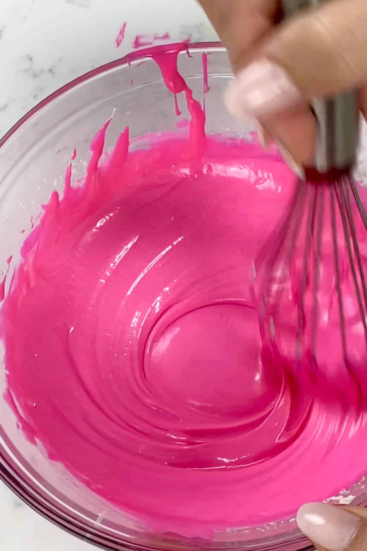 Pink water ganache in a clear bowl being mixed with a whisk.