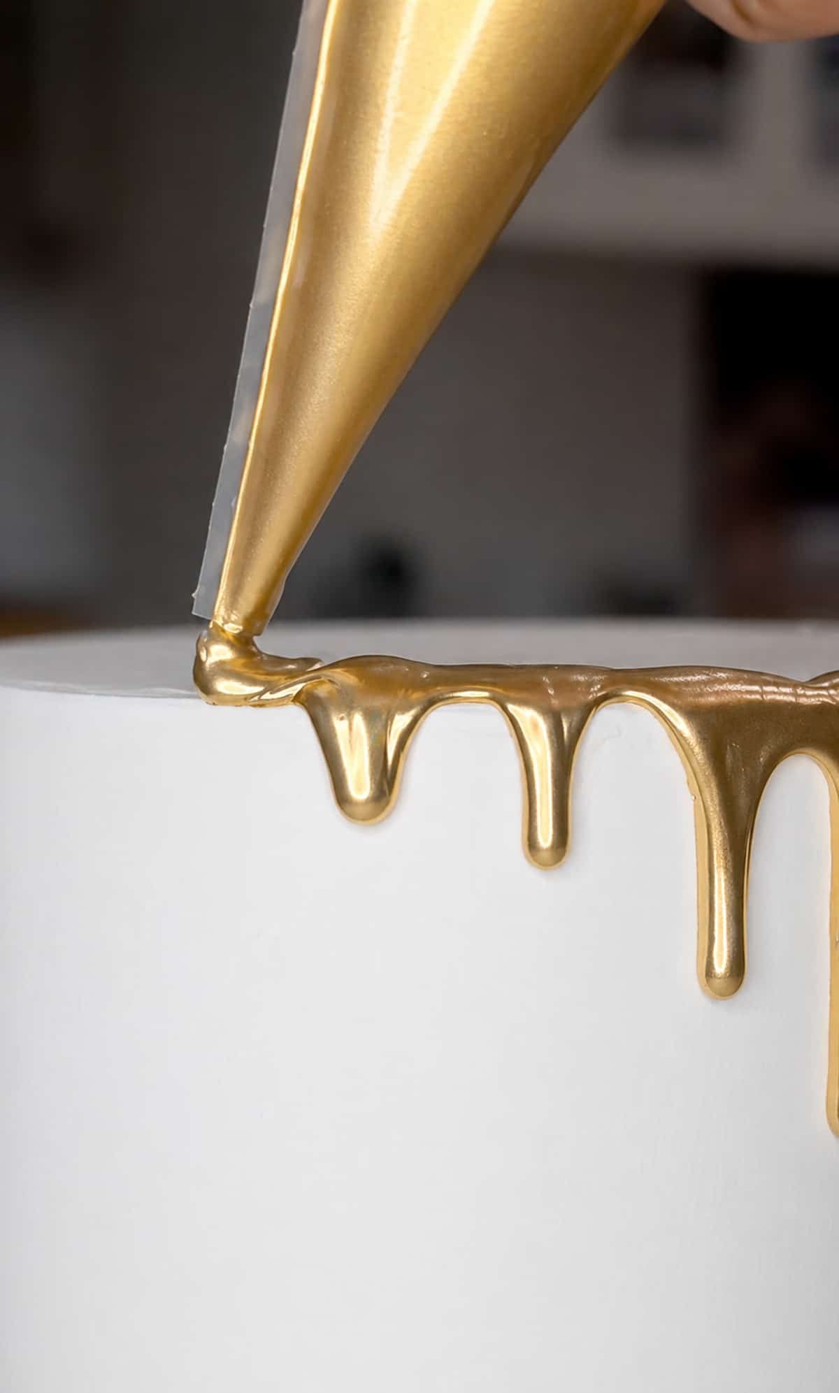 Close up of a piping bag piping gold drip onto a white cake.
