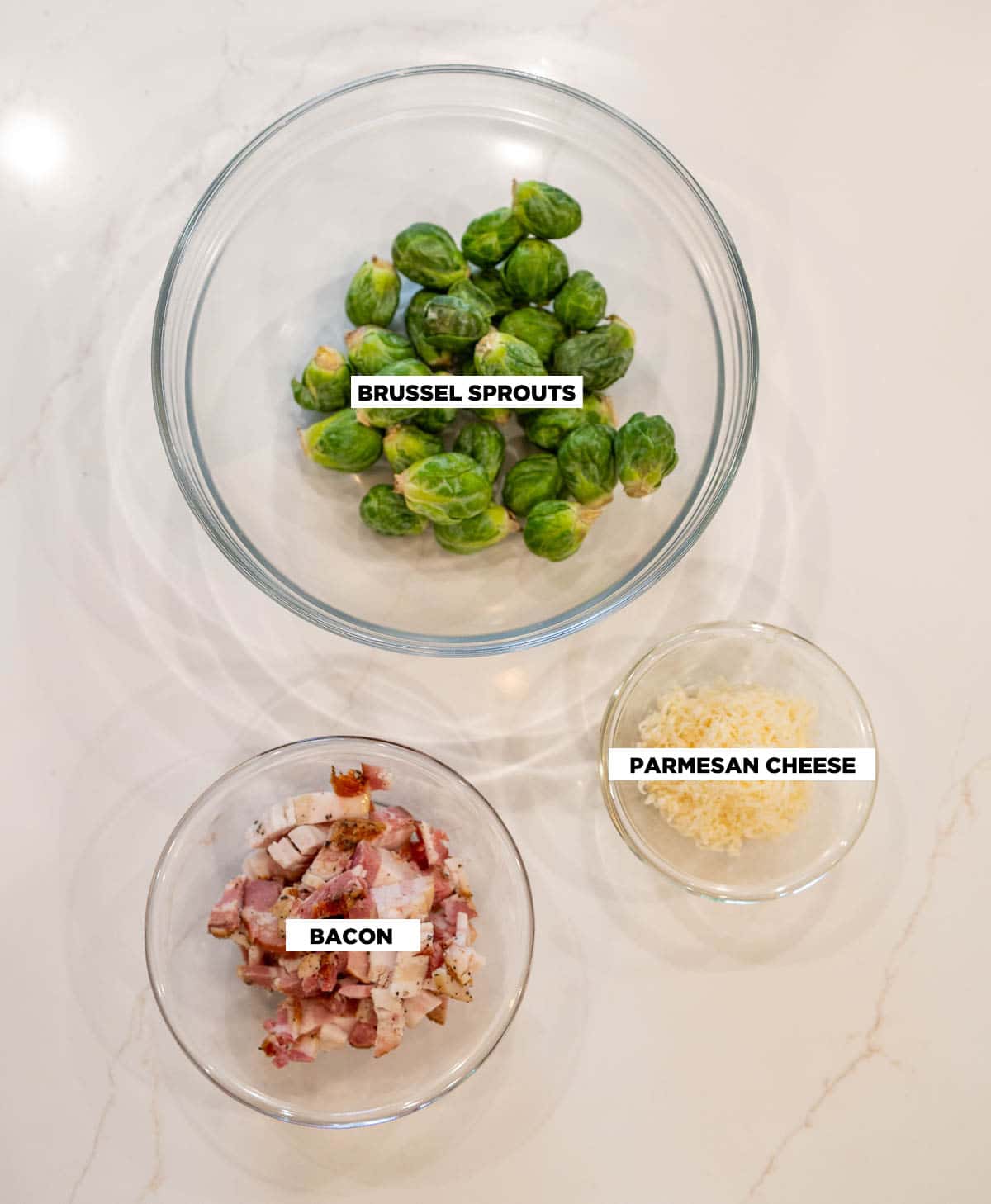 Ingredients for Bacon Brussel Sprouts, featuring: a bowl of Brussel sprouts, a bowl of bacon cut up into small pieces, and a small bowl of freshly grated Parmesan cheese. 