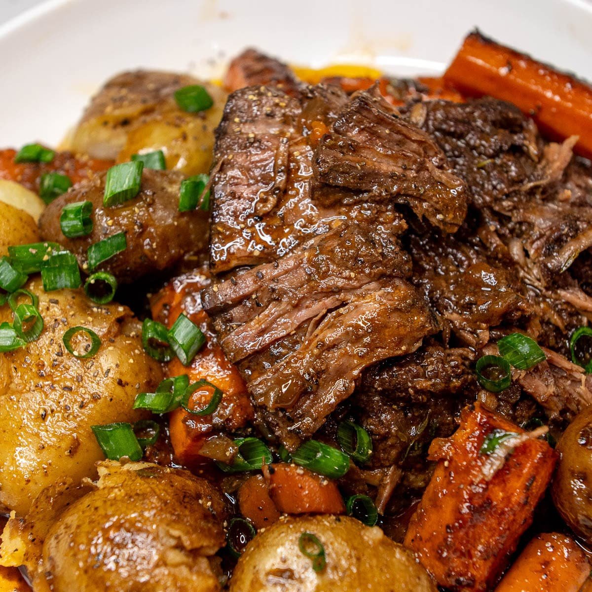 A close-up shot of a Braised Beef roast on a plate, served with roasted potatoes and carrots, and garnished with green onion. 