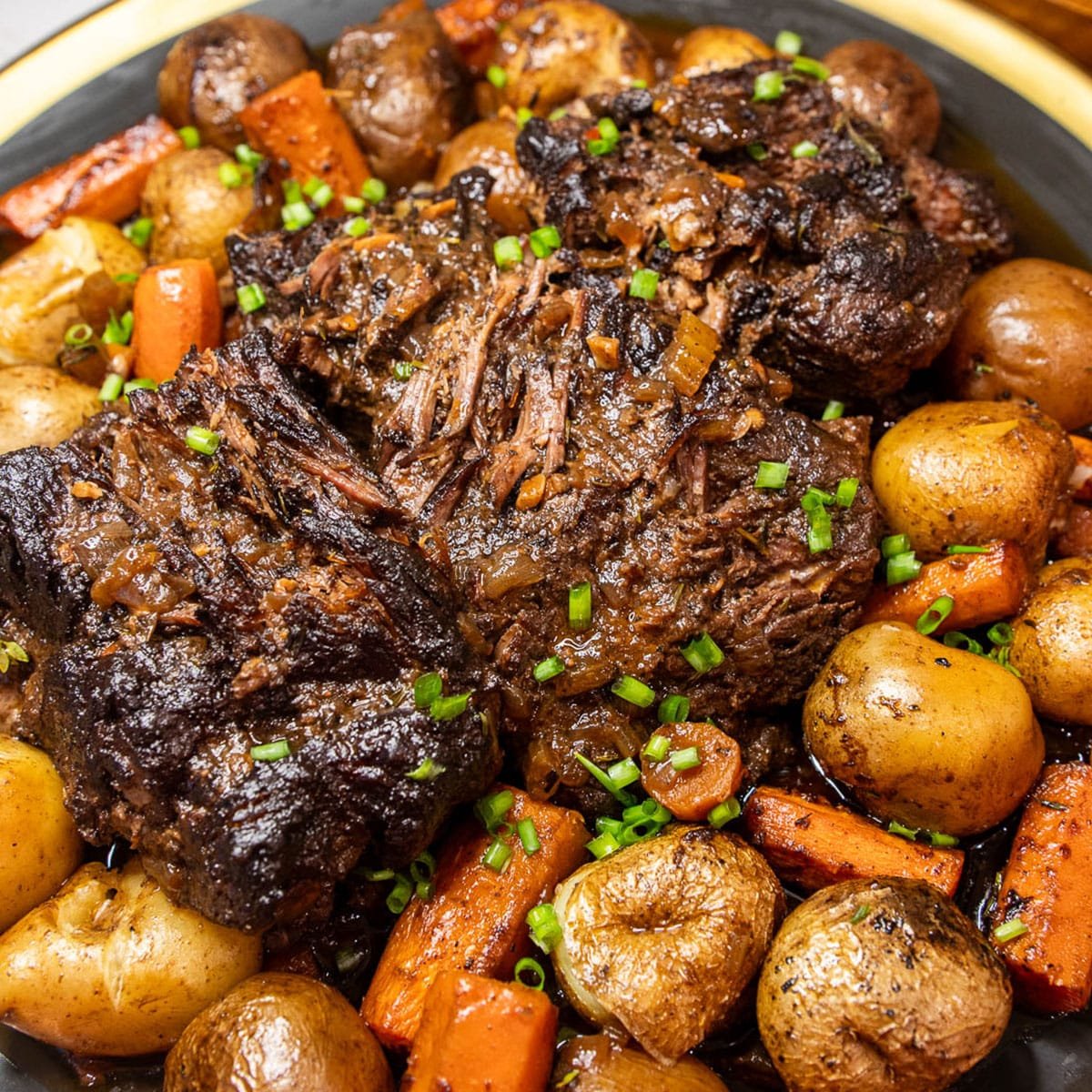 Braised Beef roast on a plate, served with roasted potatoes and carrots, and garnished with green onion. 