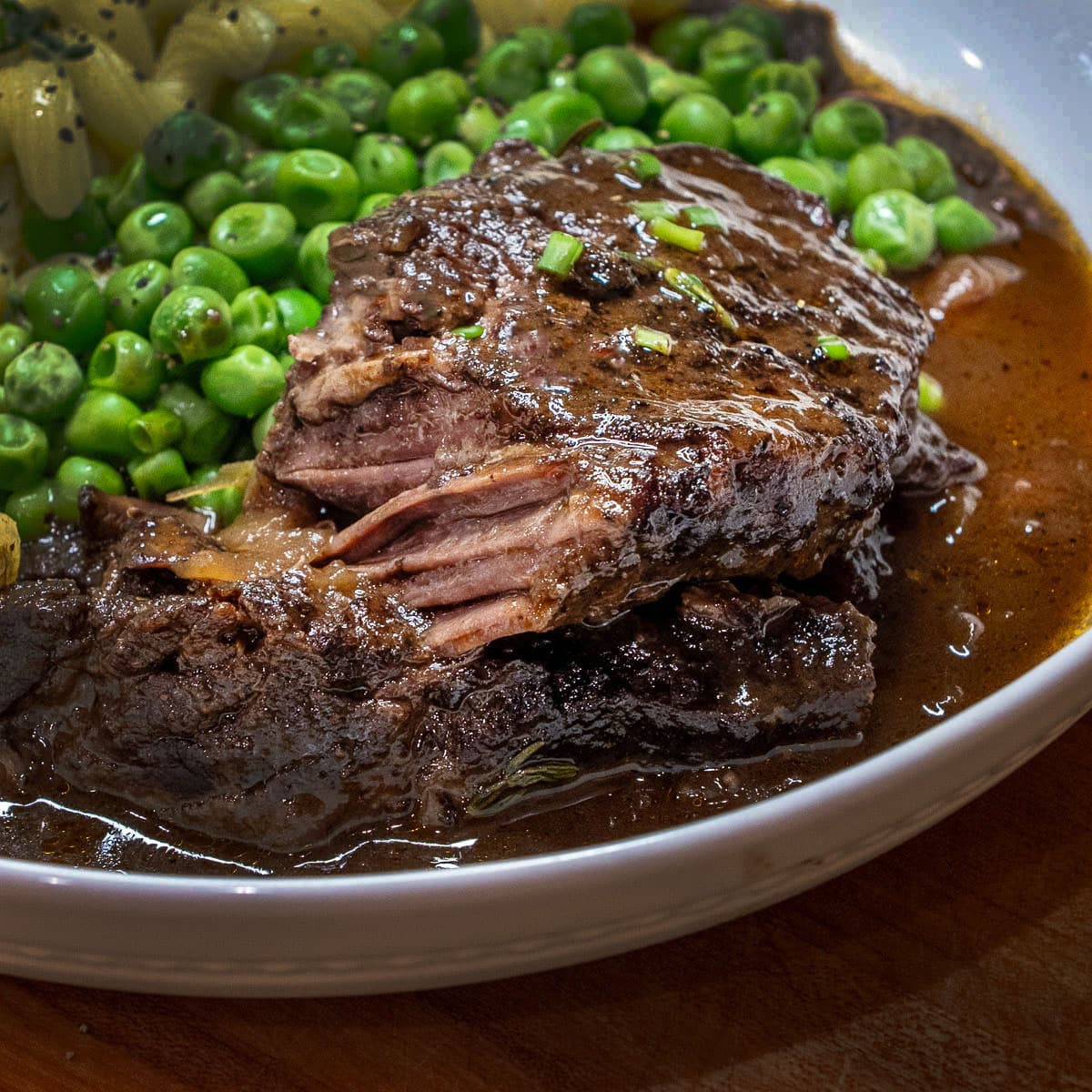 Braised Beef Short Ribs served on a plate with the red wine sauce, garnished with green onion, and a side of peas and pasta.