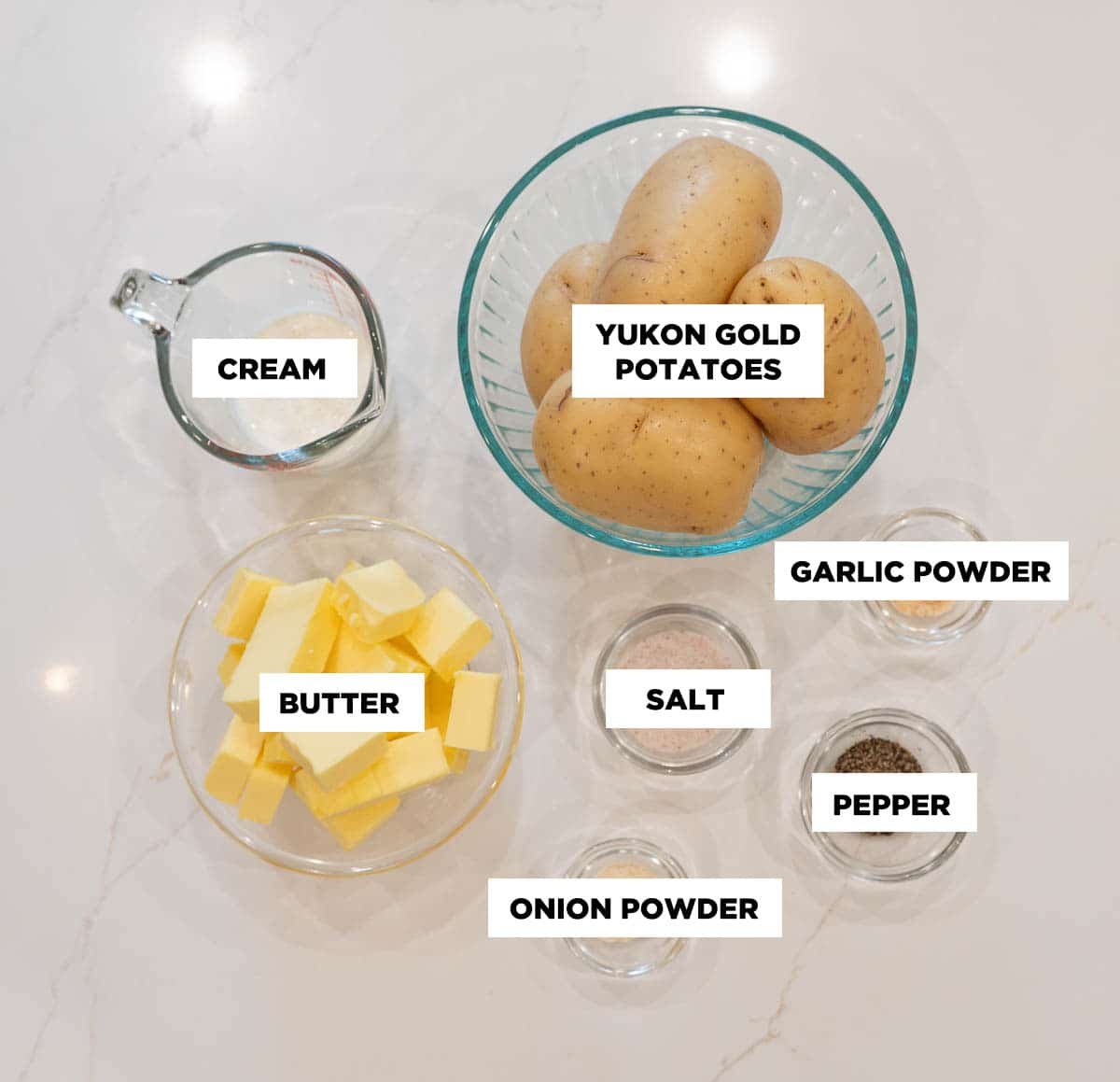 A photo of ingredients for creamy mashed potatoes featuring: heavy cream, Yukon gold potatoes, garlic powder, salt, pepper, and onion powder.