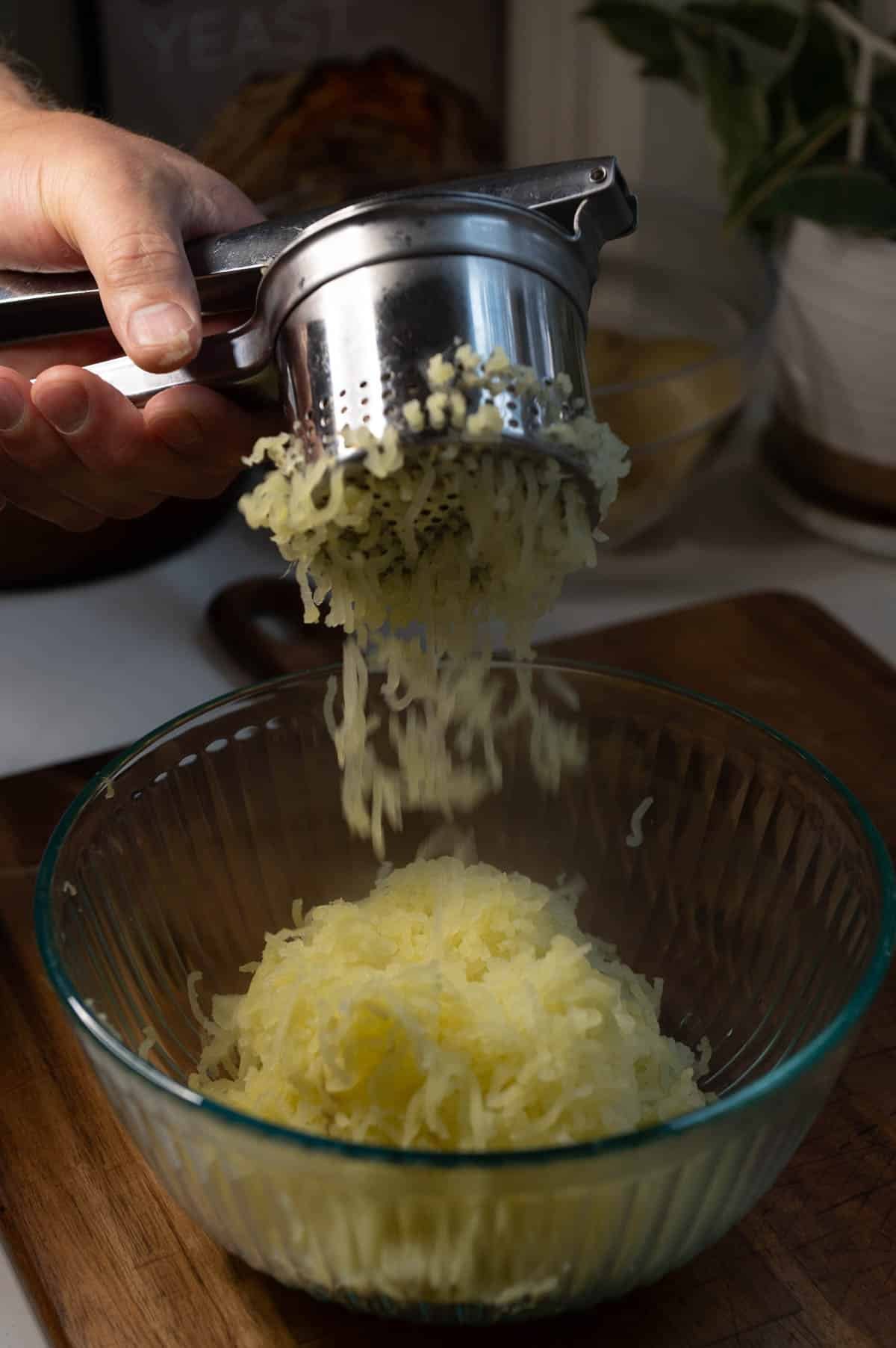 A hand holding a stainless steel potato ricer, which is pressing the potatoes out and into a glass bowl.