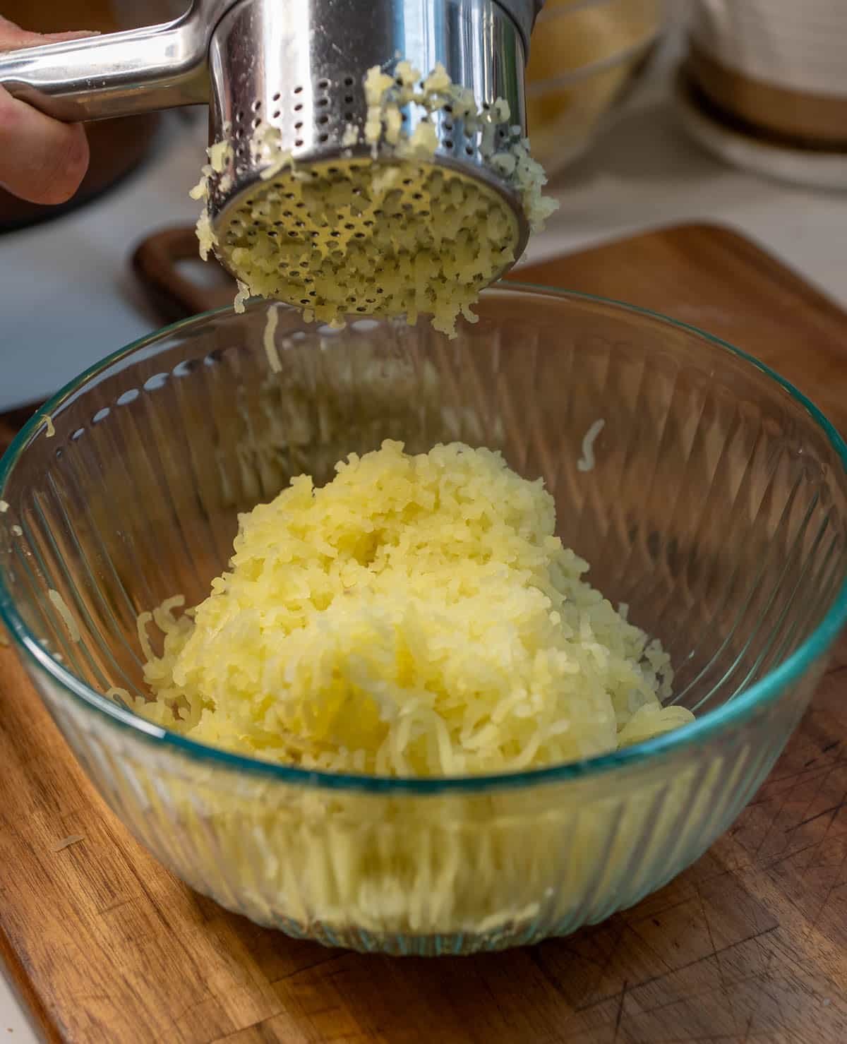 A potato ricer pressing potatoes out into fine pieces and into a glass bowl.