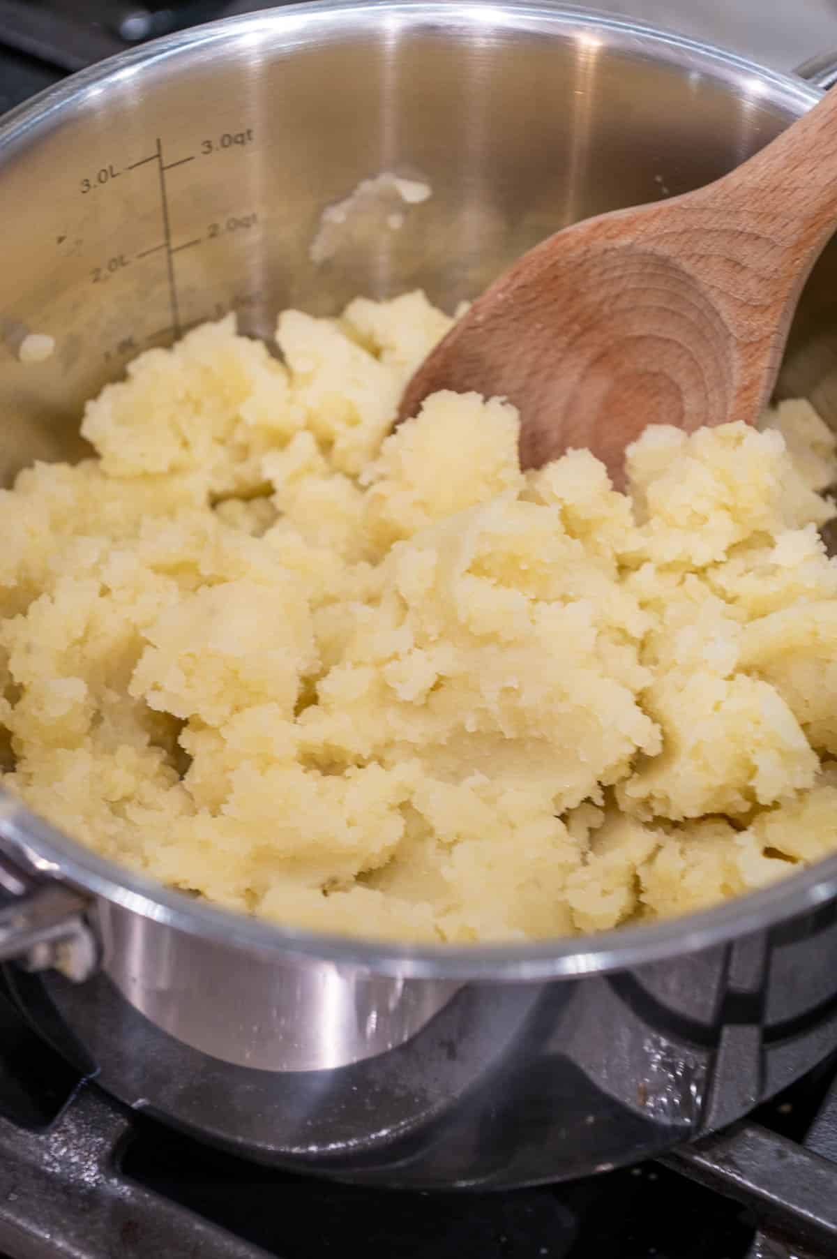 Mashed potatoes in a large cooking pot with a wooden spoon mixing it.