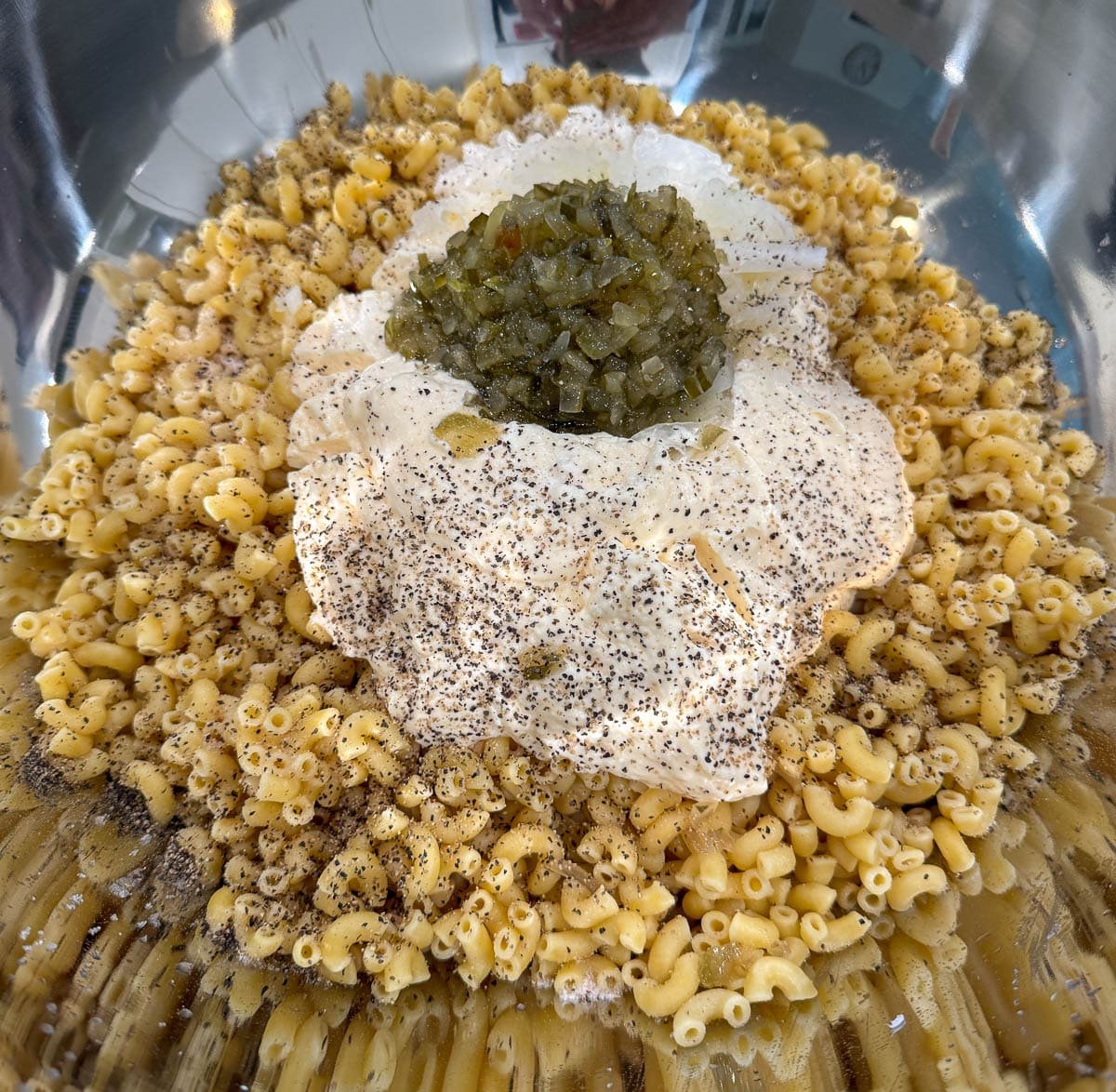Cooked macaroni in a large bowl topped with mayo, pickles, pickle juice, and seasoning. 