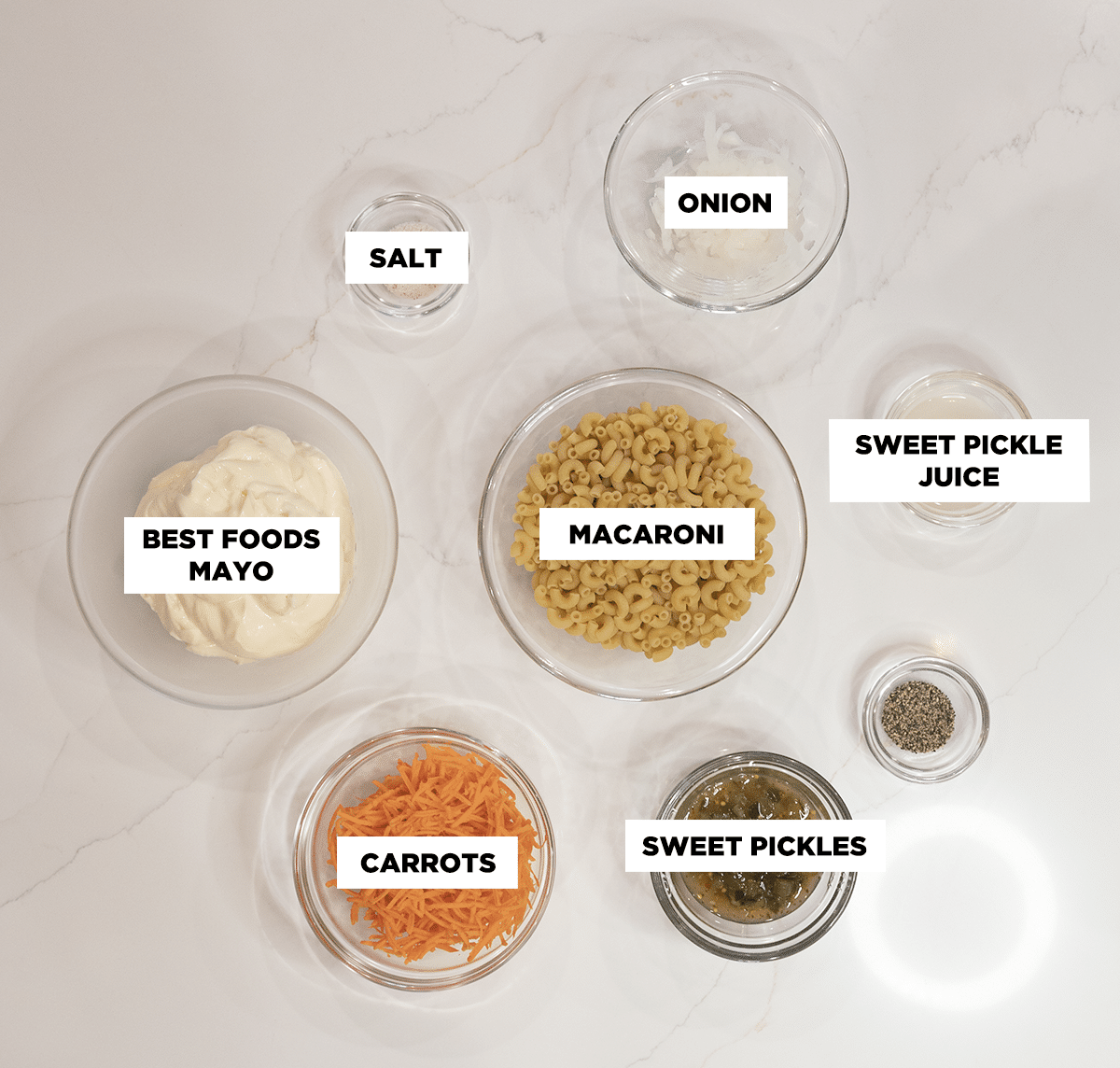 Photo of ingredients to make Hawaiian Mac Salad, featuring bowls of: salt, onion, sweet pickle juice, sweet pickles, pepper, carrots, dry macaroni noodles, and Best Foods mayo.