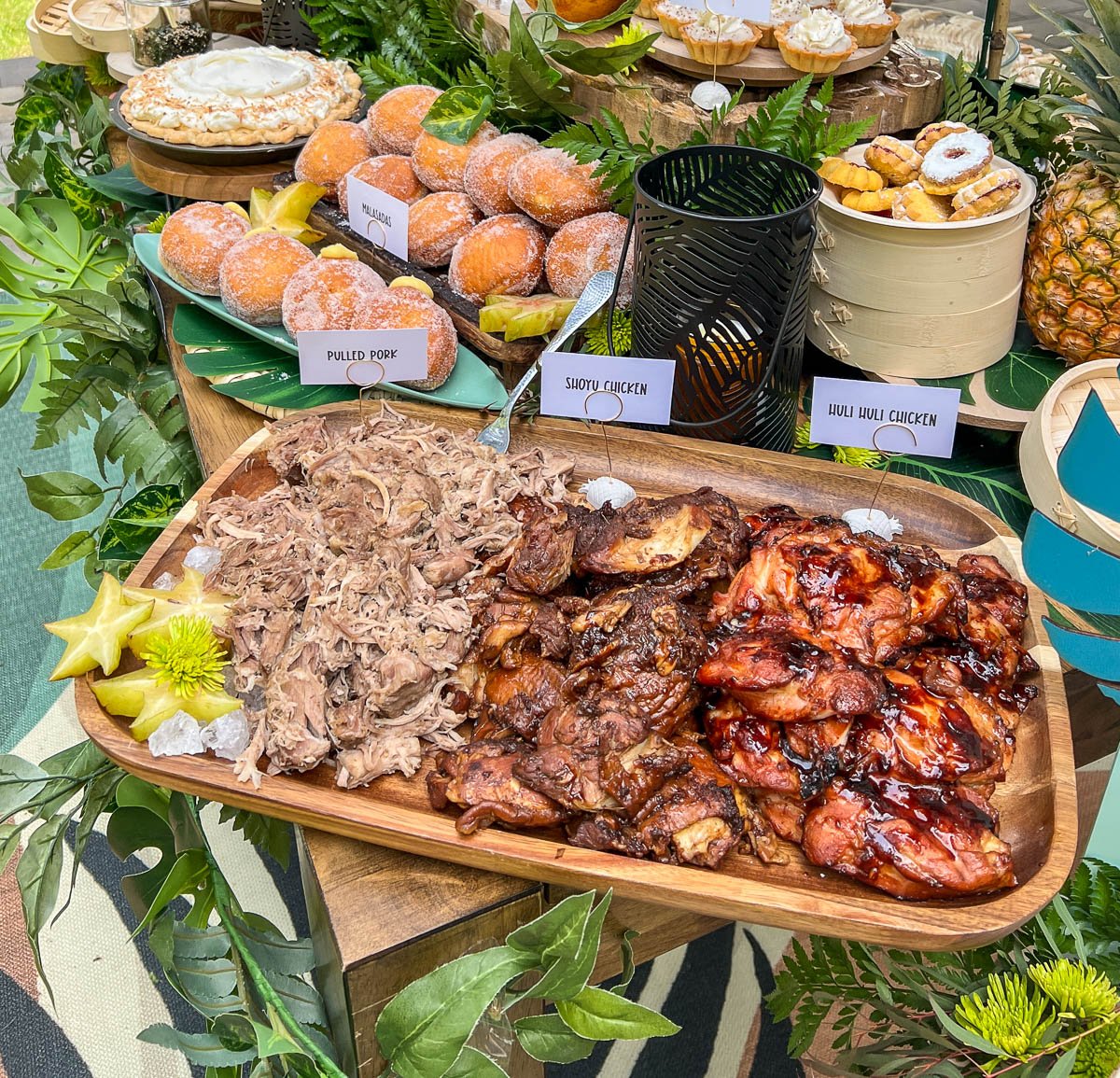 A whole Hawaiian feast on a decorative, Island-themed table with  a wooden tray of the Kalua Pork, Shoyu Chicken, and Huli Huli Chicken.