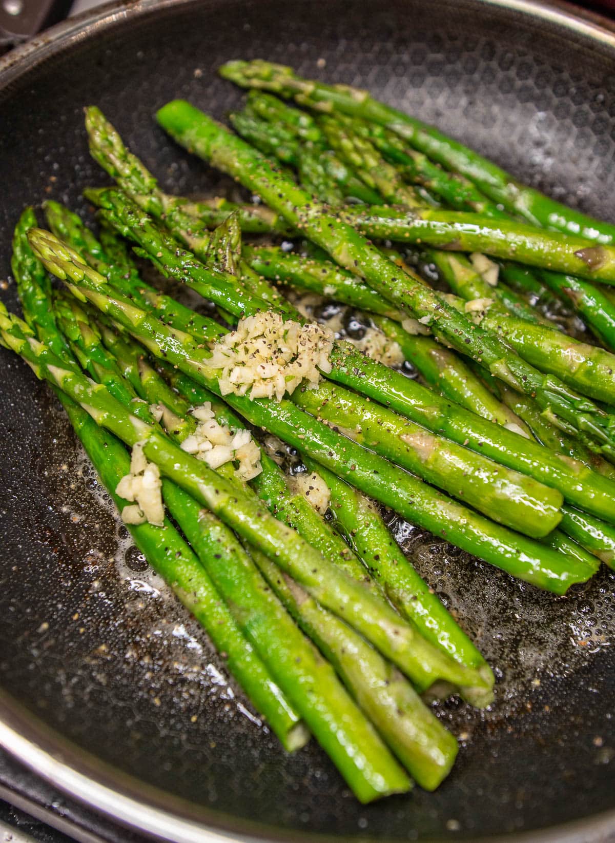 Asparagus sautéing in a skillet with freshly minced garlic on top and seasoned with salt and pepper. 