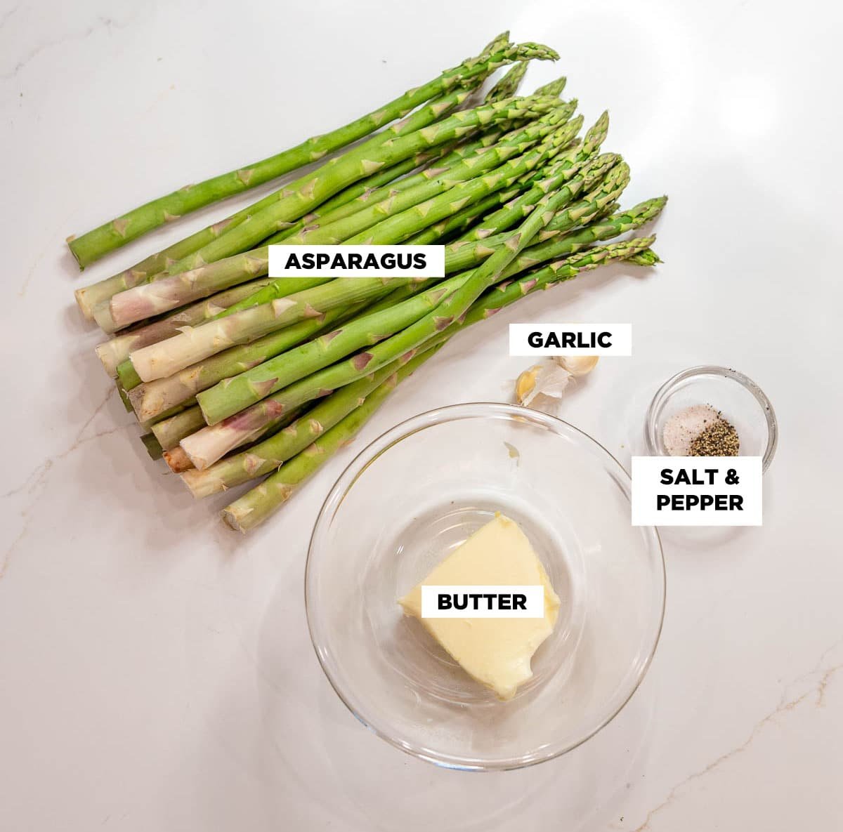 Ingredient shot for the Sautéed Asparagus recipe, featuring: a bunch of raw asparagus, two garlic cloves, a small bowl of salt and pepper, and a bowl of butter. 