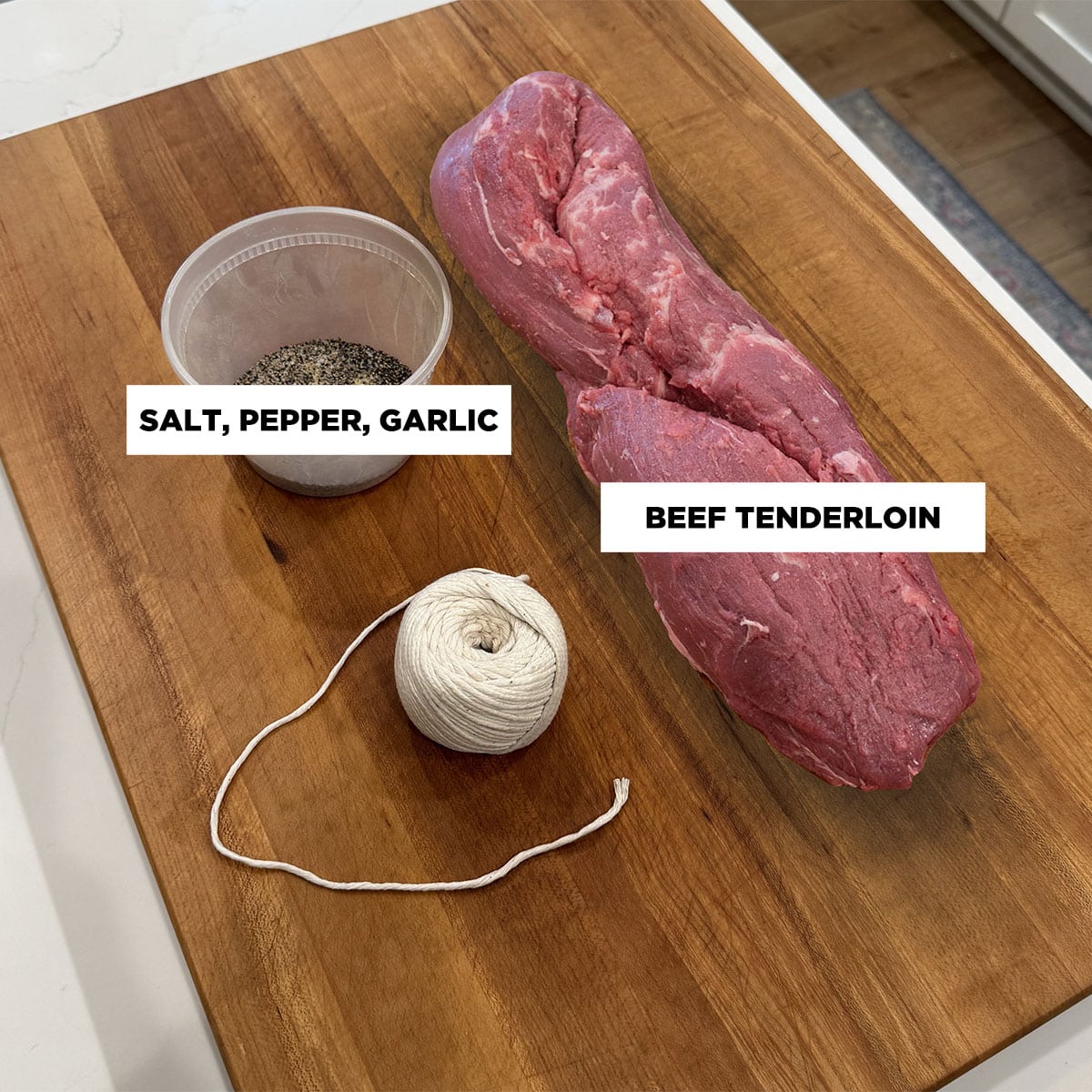 A photo of ingredients for smoked beef tenderloin, featuring: a whole, raw beef tenderloin, butcher's twine, and a plastic container of salt, pepper, and granulated garlic.