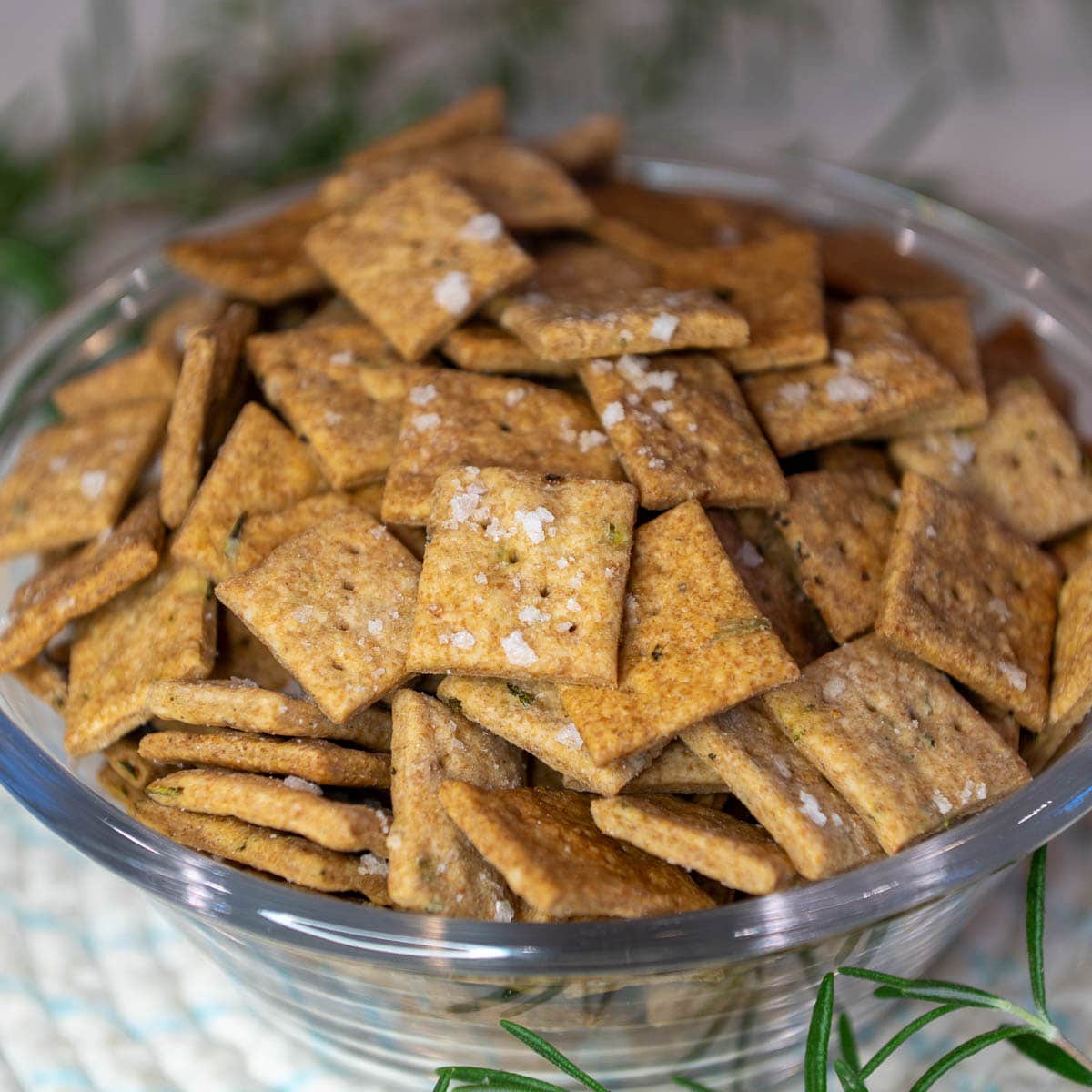 A close-up shot of sourdough discard crackers topped with flaky salt in a glass bowl with fresh rosemary on the table.