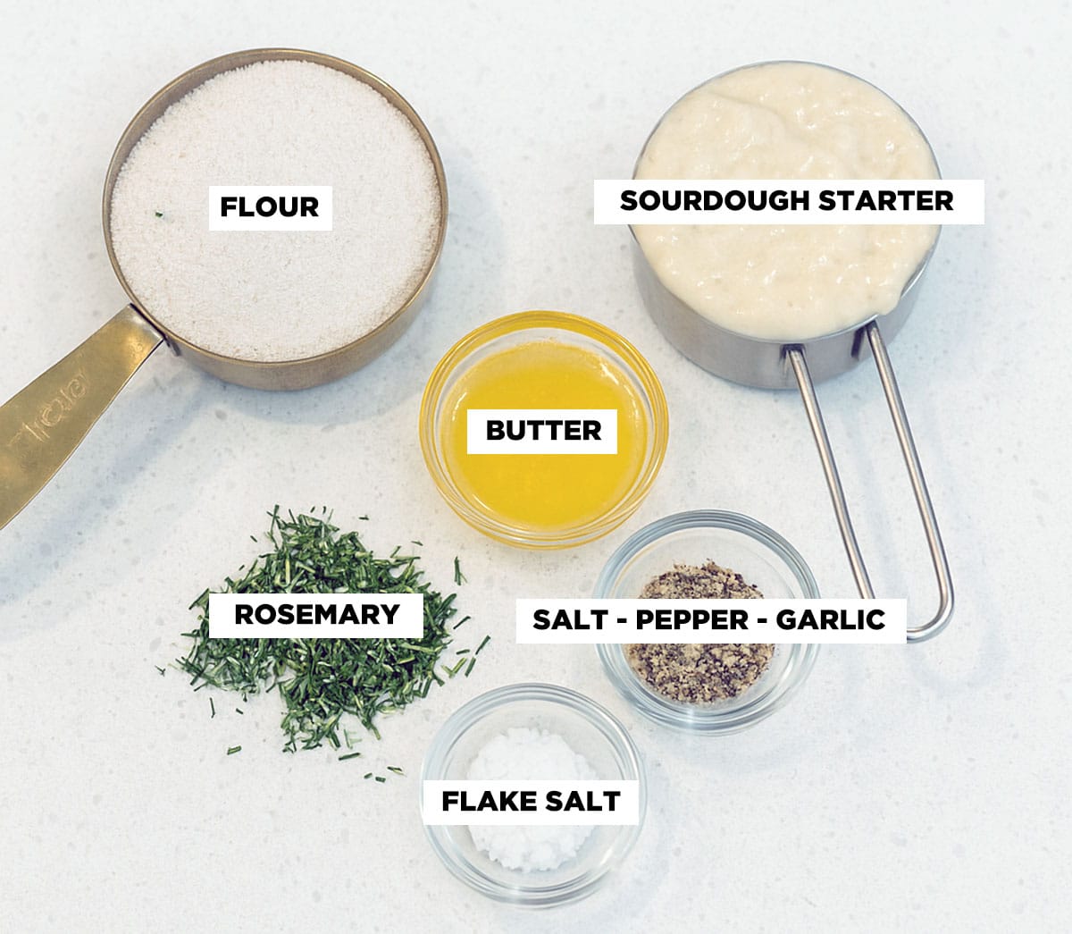 An ingredient shot featuring flour, sourdough starter, melted butter, fresh rosemary, flake salt, and a small bowl of salt, pepper, and garlic powder.