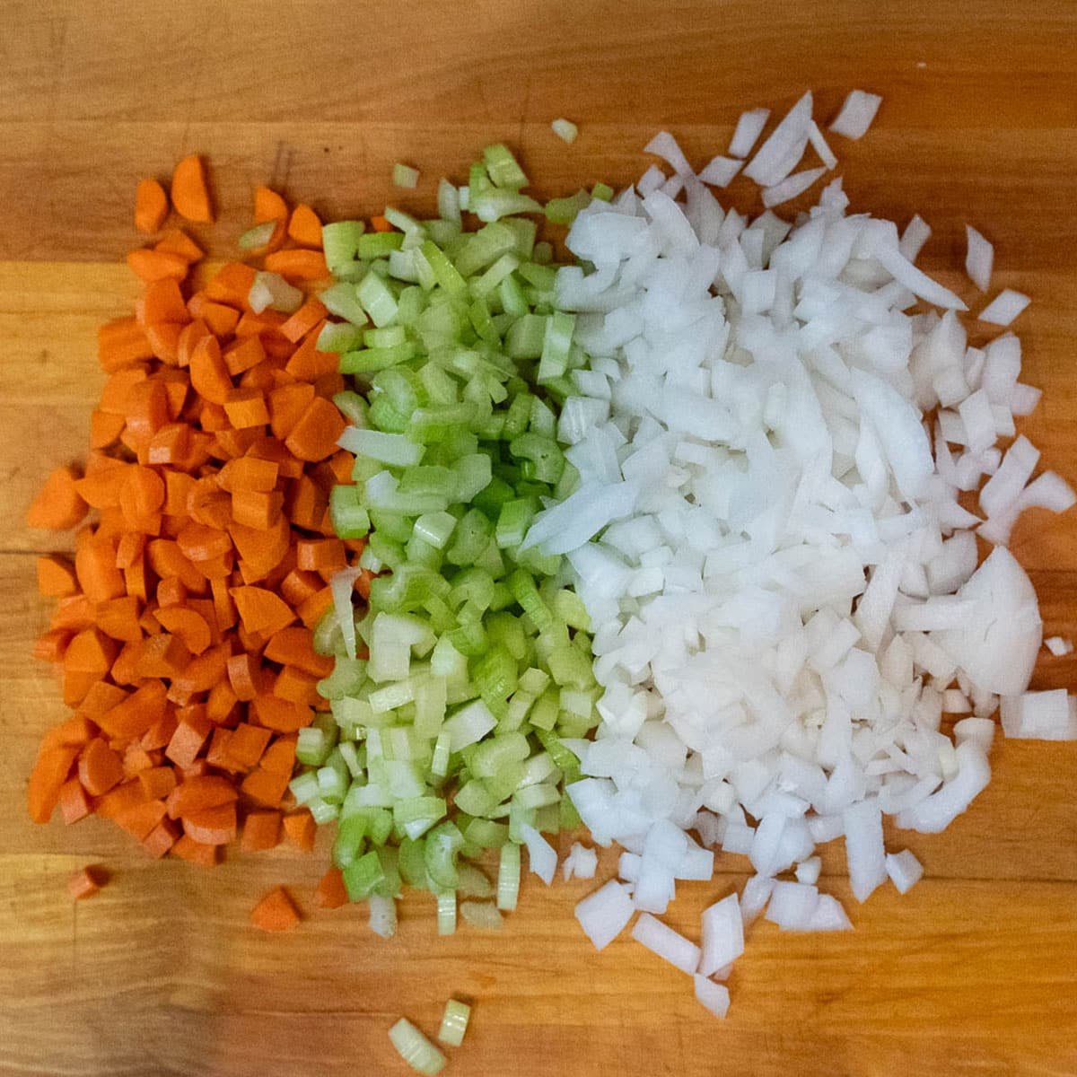 Chopped carrots, celery, and onion in a 2:1:1 ratio, grouped together, side-by-side on a wooden cutting board. 