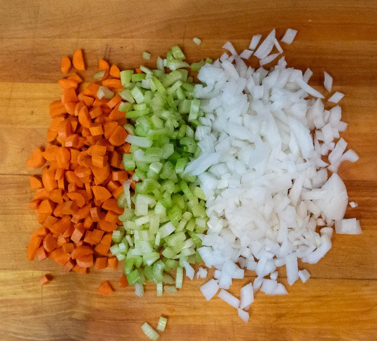 Chopped carrots, celery, and onion in a 2:1:1 ratio, grouped together, side-by-side on a wooden cutting board. 