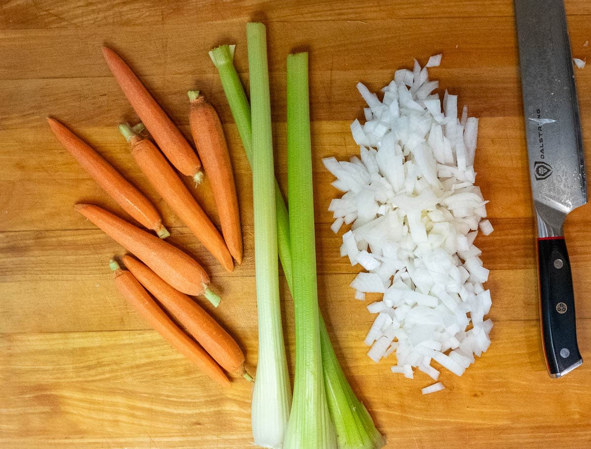 Ingredients for the Mirepoix recipe: Diced onion, three celery stalks, and seven small peeled carrots on a wooden cutting board.
