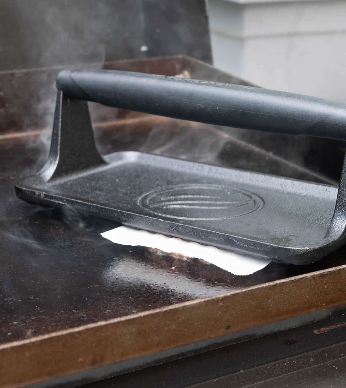 A blackstone burger press is smashing the burger patty on a hot griddle with parchment paper in between the burger press and the burger patty.