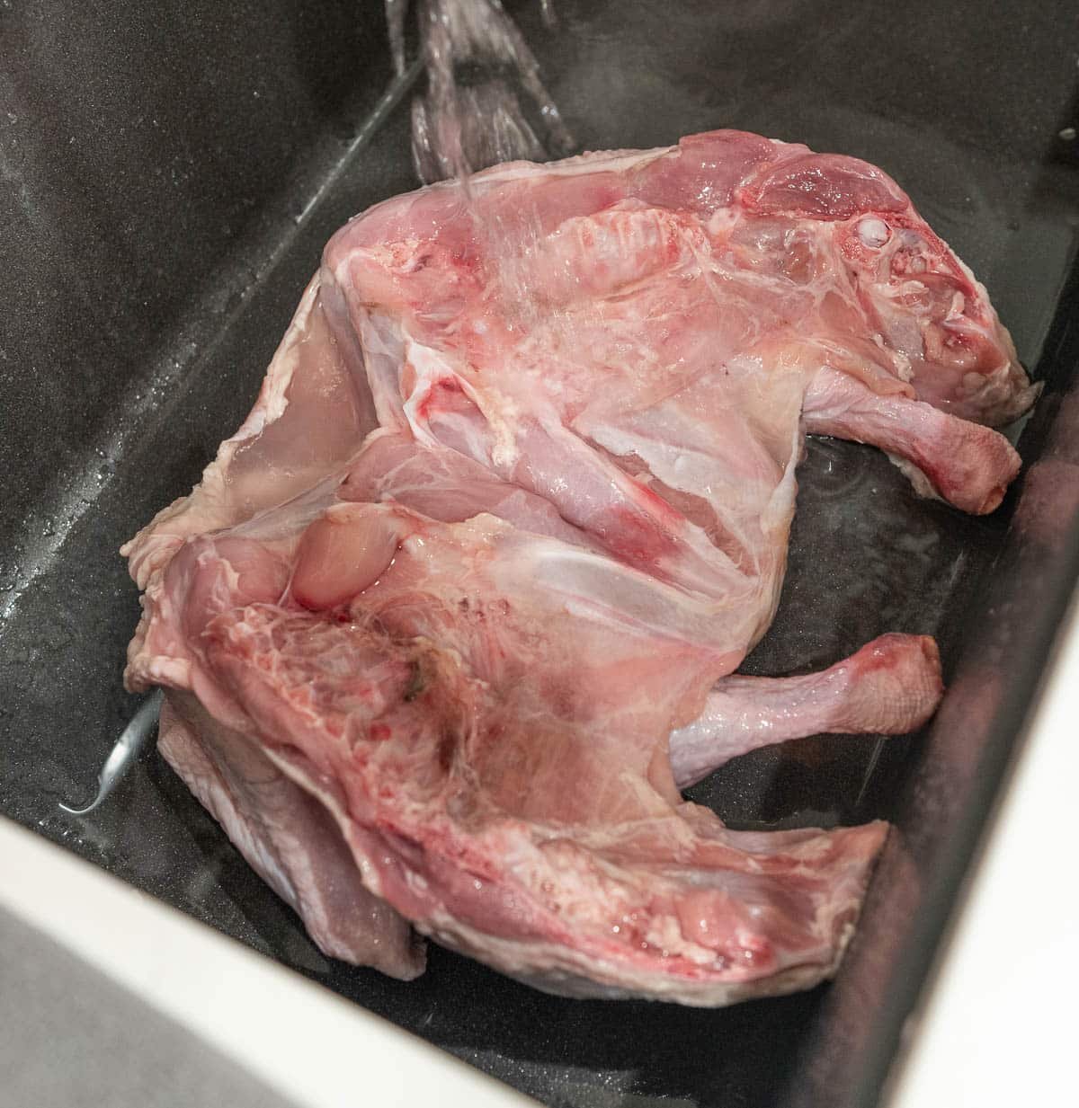 A raw turkey is being rinsed in the sink with running water. 
