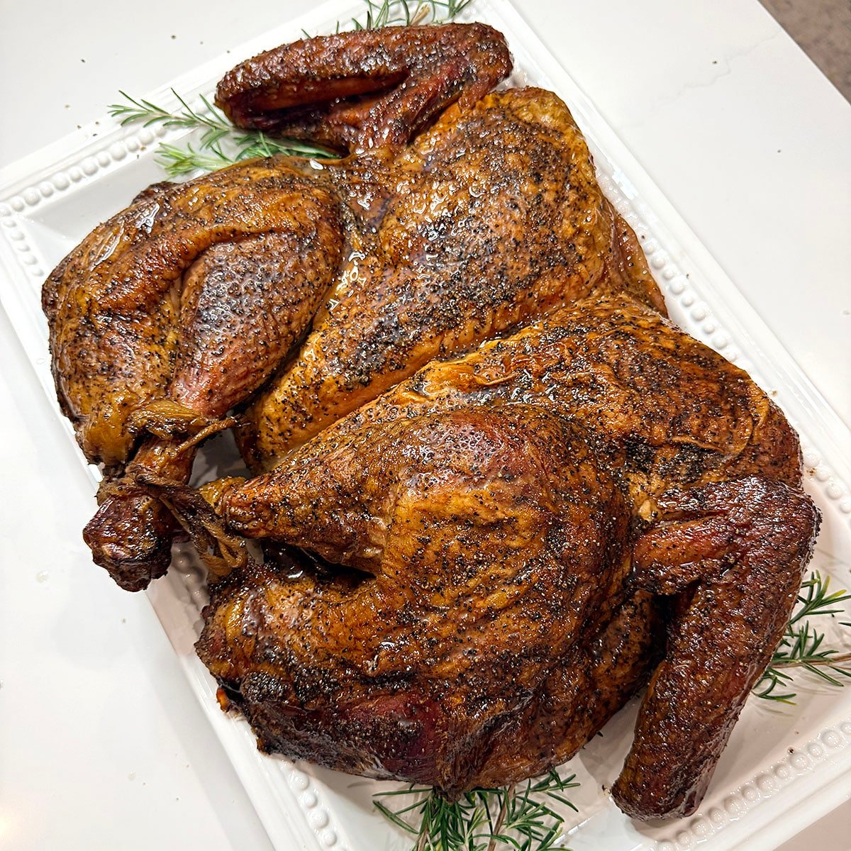 A whole, spatchcocked smoked turkey on a white platter with fresh herbs. 