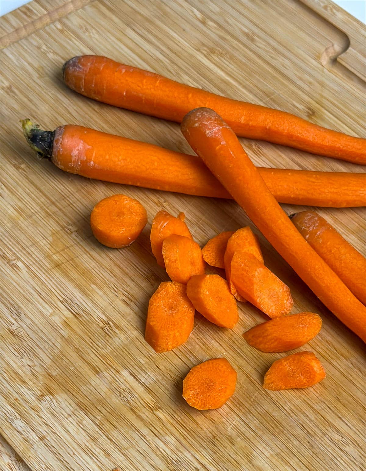 Peeled and washed carrots next to a pile of carrots cut into pieces on a wooden cutting board.