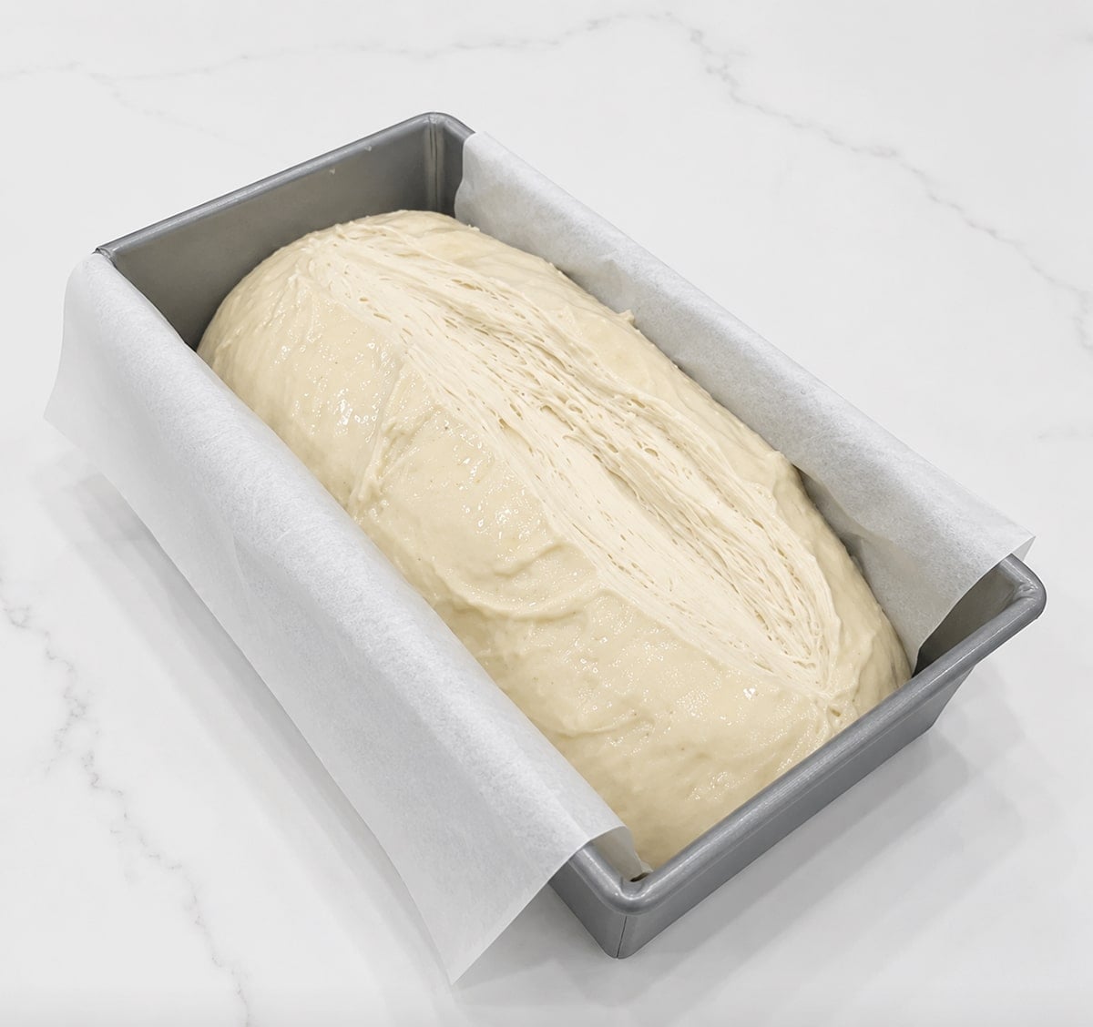 Loaf of scored dough in a loaf pan with parchment paper.