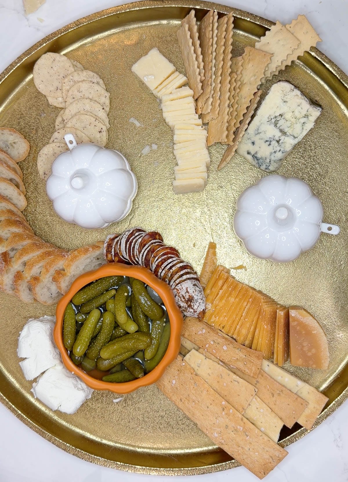 A gold tray with cut cheeses, bread, crackers, and pickles on it.