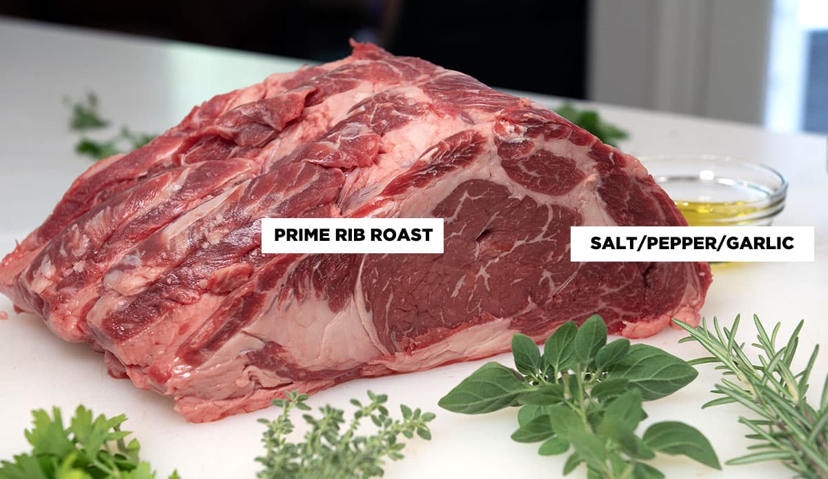 A raw prime rib roast with fresh herbs and a salt glass bowl of oil in the background.