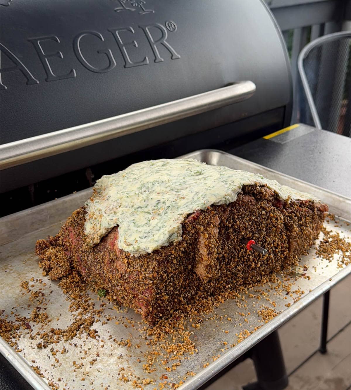 A whole raw and seasoned prime rib with a butter and herb topping on a sheet pan in front of the smoker.