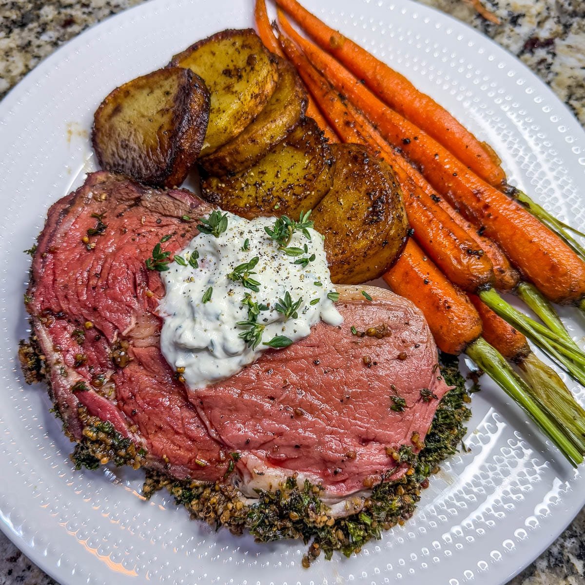 A close-up photo of a slice of smoked prime rib with sauce, a side or roasted potatoes and carrots on a white plate.