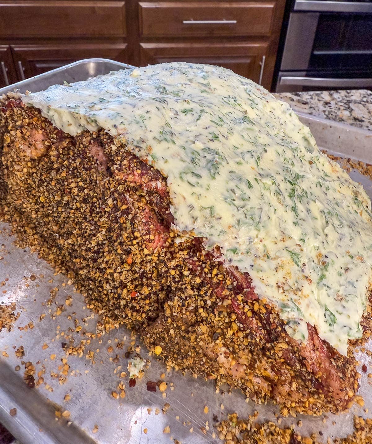 A whole raw and seasoned prime rib with a butter and herb topping on a sheet pan.