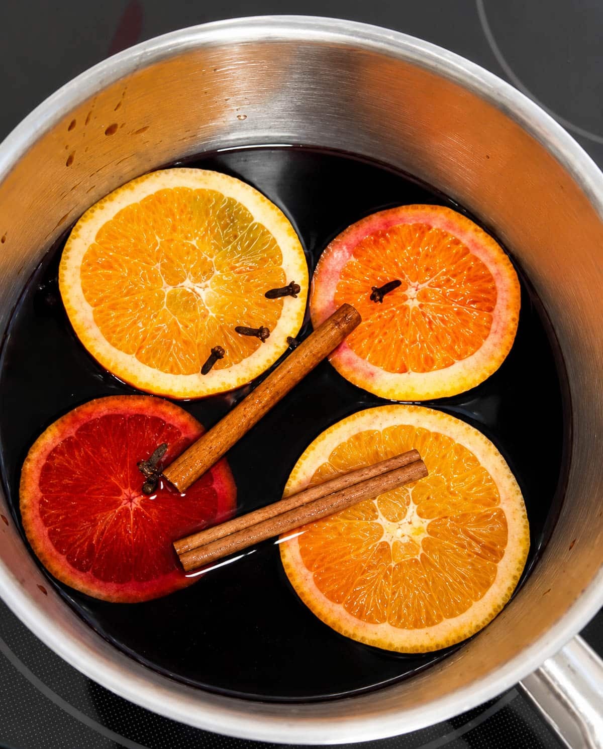 Slices of citrus, cinnamon sticks, and cloves with wine in a saucepan.