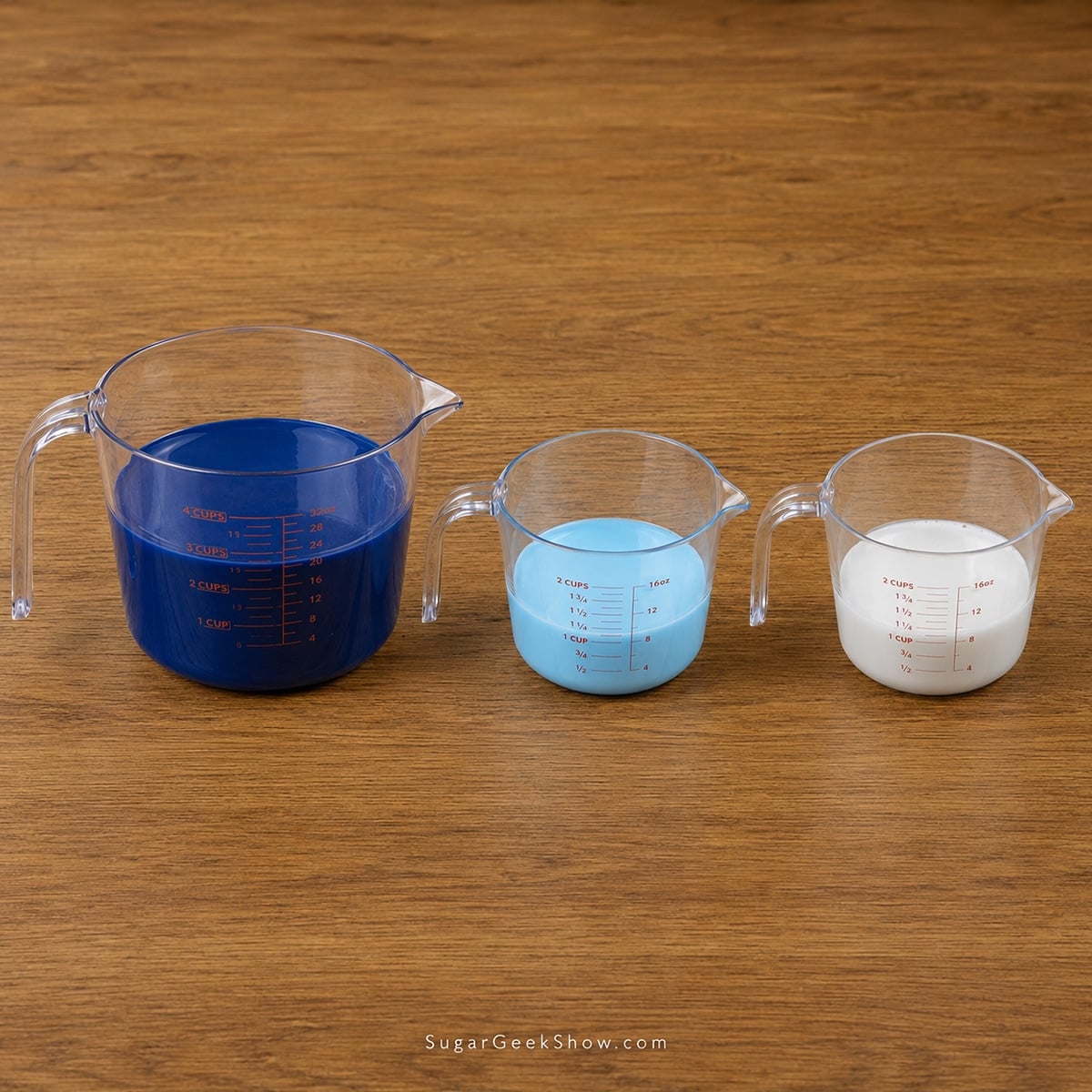 Three measuring cups with blue, light blue and white mirror glaze in them on a wooden countertop.