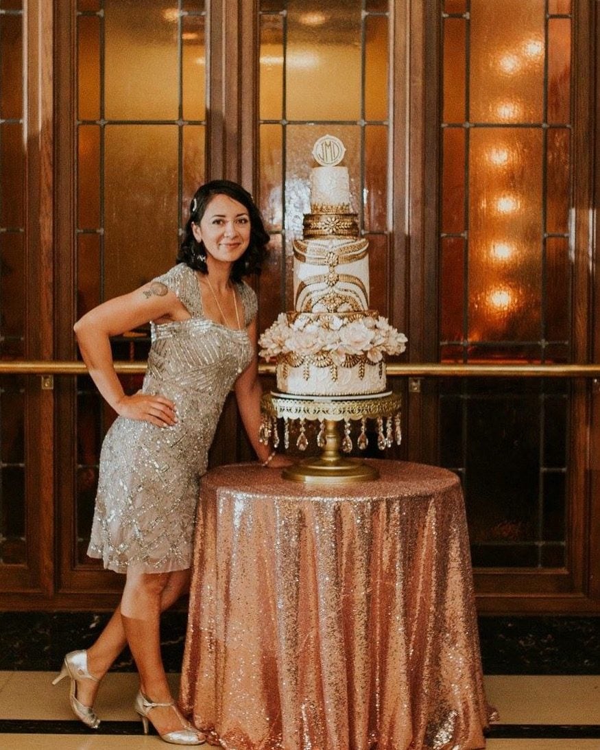 Liz Marek beside a three-tier Great Gatsby themed wedding cake with gold art deco piping, sugar flowers, and a monogram topper on a crystal cake stand