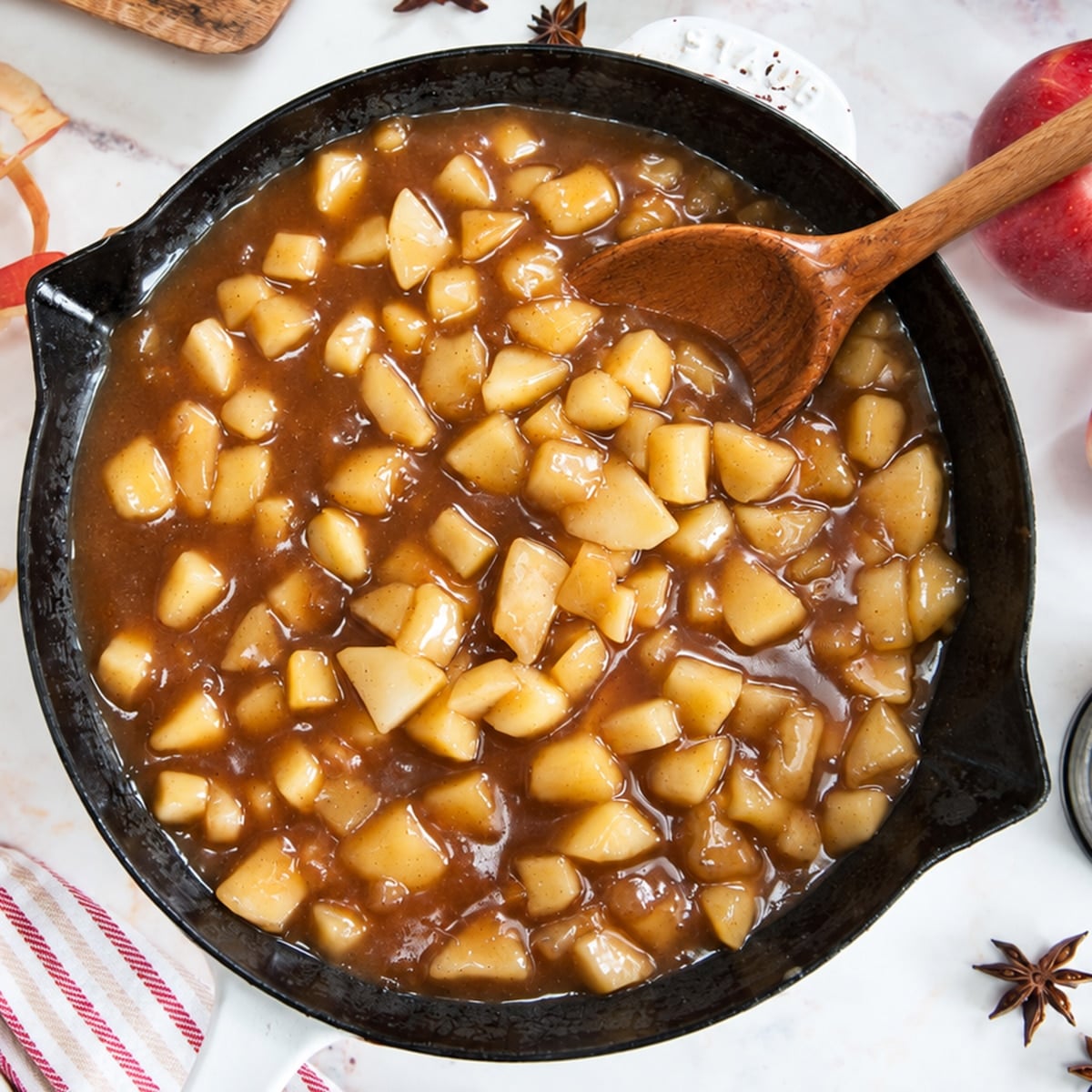 Homemade apple pie filling with cubed apples in a cast iron skillet with a wooden spoon, surrounded by fresh apples