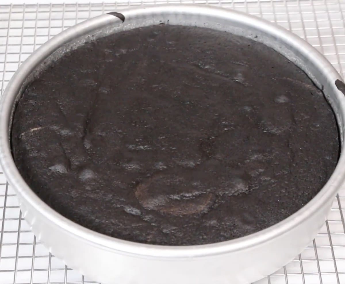 Baked black velvet cake layer in a cake pan on a cooling rack.
