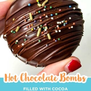 Close up of a hand holding a hot chocolate bomb drizzled with chocolate and topped with rainbow sprinkles.
