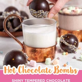 Hand lowering a shiny hot chocolate bomb with a sprinkle band into a copper mug of milk, with more bombs and mini marshmallows around it.