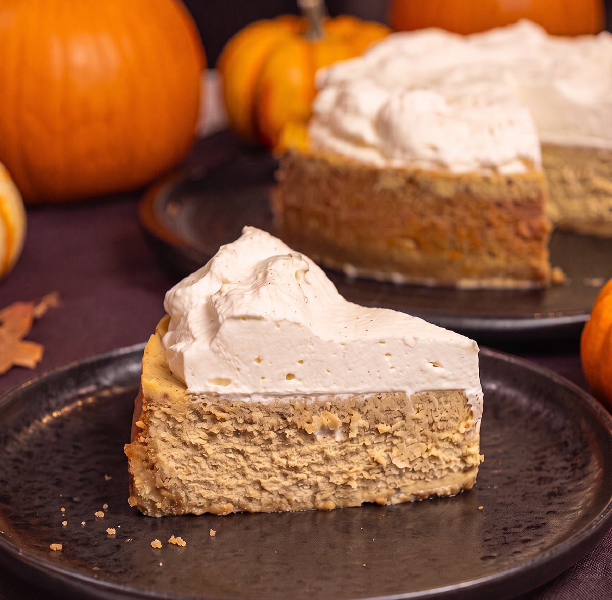 A slice of pumpkin cheesecake with whipped cream on a black plate, with pumpkins and the whole cheesecake in the background