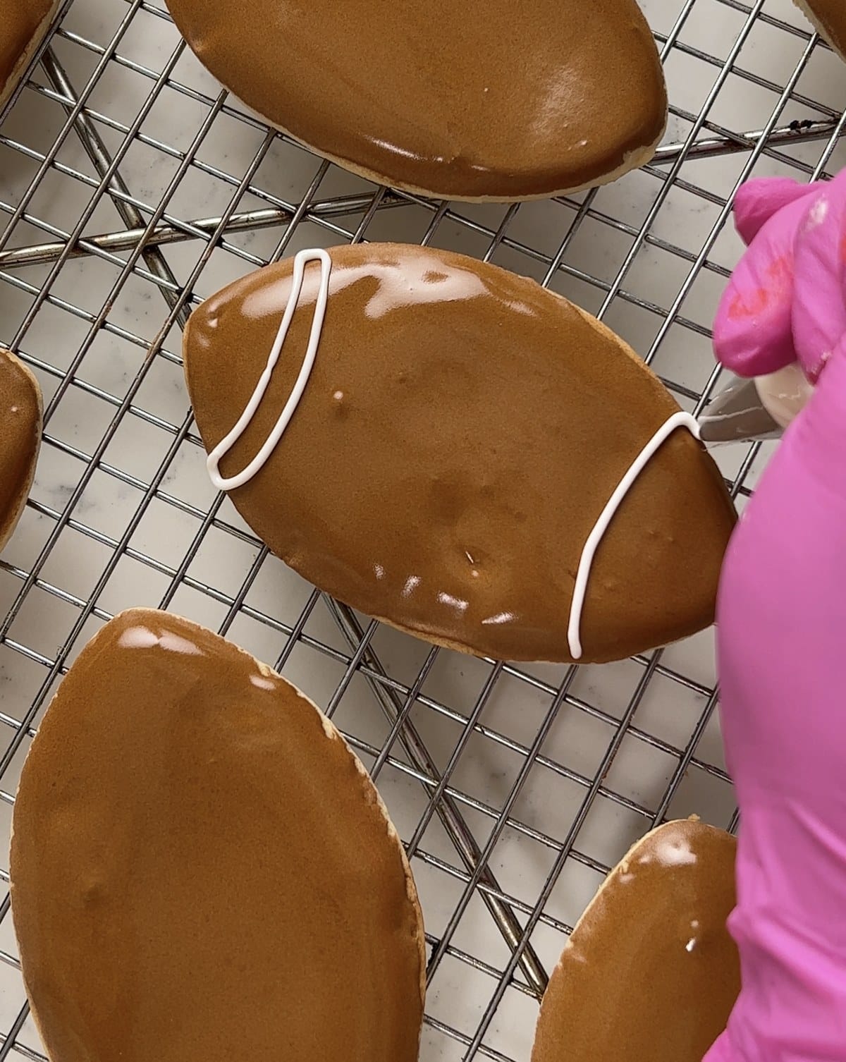 Piping white royal icing laces onto a brown football cookie.